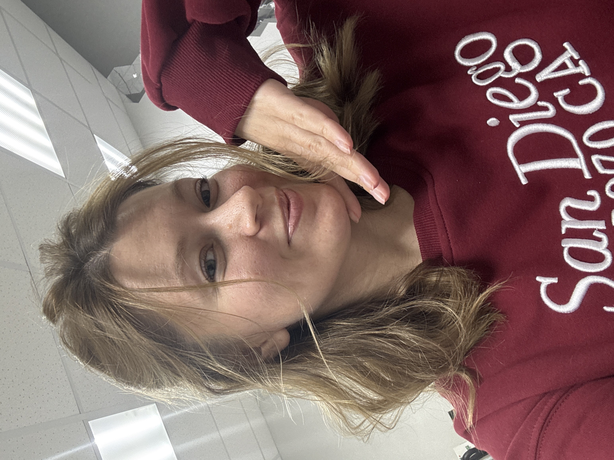 A portrait-style selfie captures a light-skinned woman with light brown, wavy hair, looking directly at the camera with a calm, pleasant expression, possibly a slight smile. She is wearing a dark red or maroon long-sleeved sweatshirt. Her left hand is raised, with fingers gently curled, touching her face near her chin or cheek.
The background suggests an indoor setting, likely an office, classroom, or commercial building. It features a white suspended ceiling with rectangular fluorescent light fixtures that are illuminated, casting bright light. Light-colored walls are visible behind her.
The woman appears to be posing for the photograph. A notable detail is the white embroidered text on her sweatshirt, which clearly reads "San Diego CA". The overall impression is one of a casual, relaxed moment. While the image is provided with the location context of Chaikovsky, Russia, there are no specific visual cues within the image itself to confirm this geographical detail.