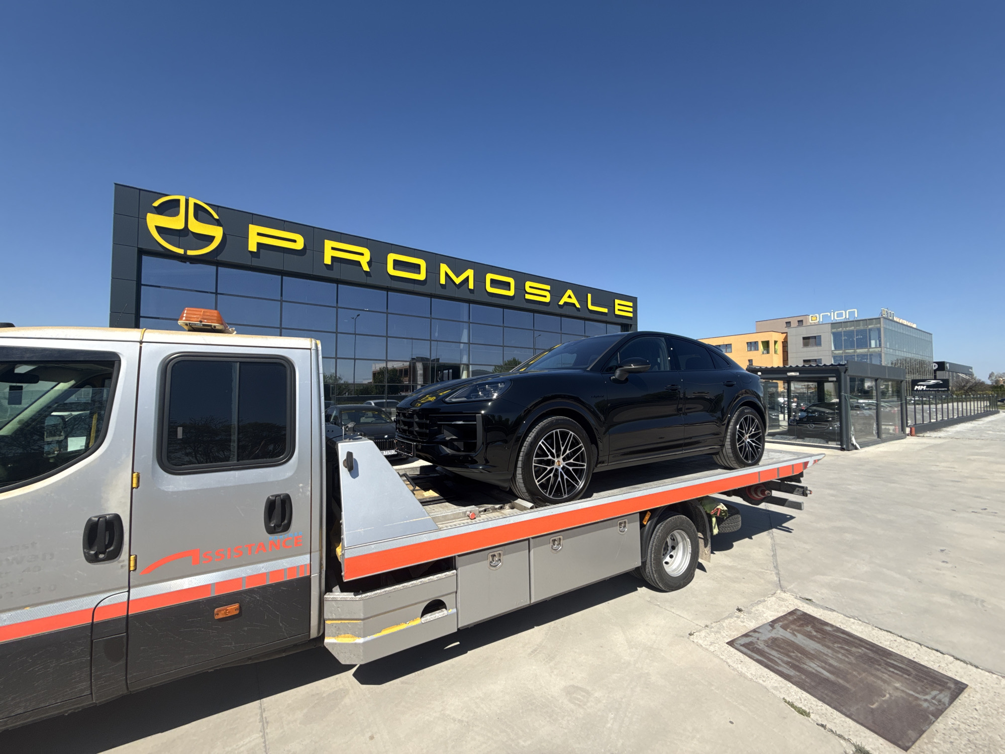 The image shows a black Porsche Cayenne Coupe being transported on a silver tow truck. The truck is parked in front of a building with the word "PROMOSALE" in yellow letters on its facade. In the background, another building with the name "Orion" can be seen. The location is Plovdiv, Bulgaria, judging by the Promosale dealership and architecture, but this information has been gathered independently. The sky is clear and blue.