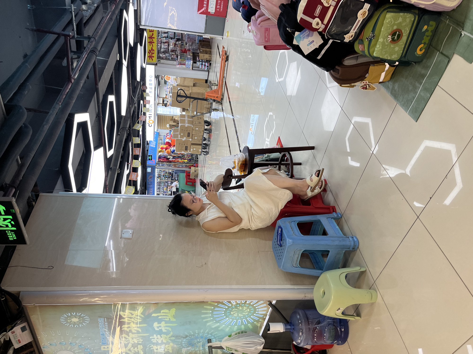 In the Liwan District of China, a woman with her hair pulled back and wearing glasses sits on a red stool, looking at her phone. She is wearing a light-colored sleeveless dress and sandals. Next to her, a blue plastic stool and a green plastic stool are visible. A large blue water dispenser is also present. In the background, a busy market scene unfolds, with various stalls displaying goods, shelves stacked with boxes, and shopping carts. The flooring is tiled, reflecting the overhead lighting fixtures, which are hexagonal and white. The scene appears to be indoors, possibly in a shopping mall or market. Several backpacks are stacked on the right side of the image.