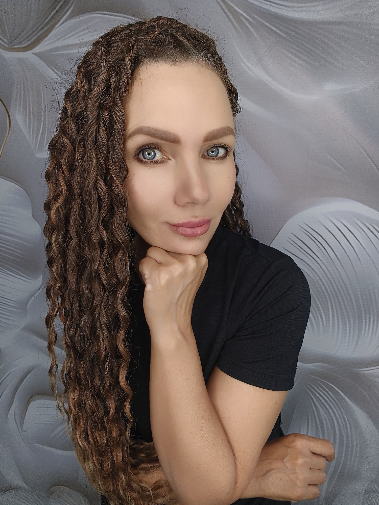 A woman with long, curly brown hair and blue eyes is shown from the chest up. She is wearing a black t-shirt and has her chin resting on her fist. She is looking directly at the camera. The background is a light gray wall with a textured pattern of abstract shapes.