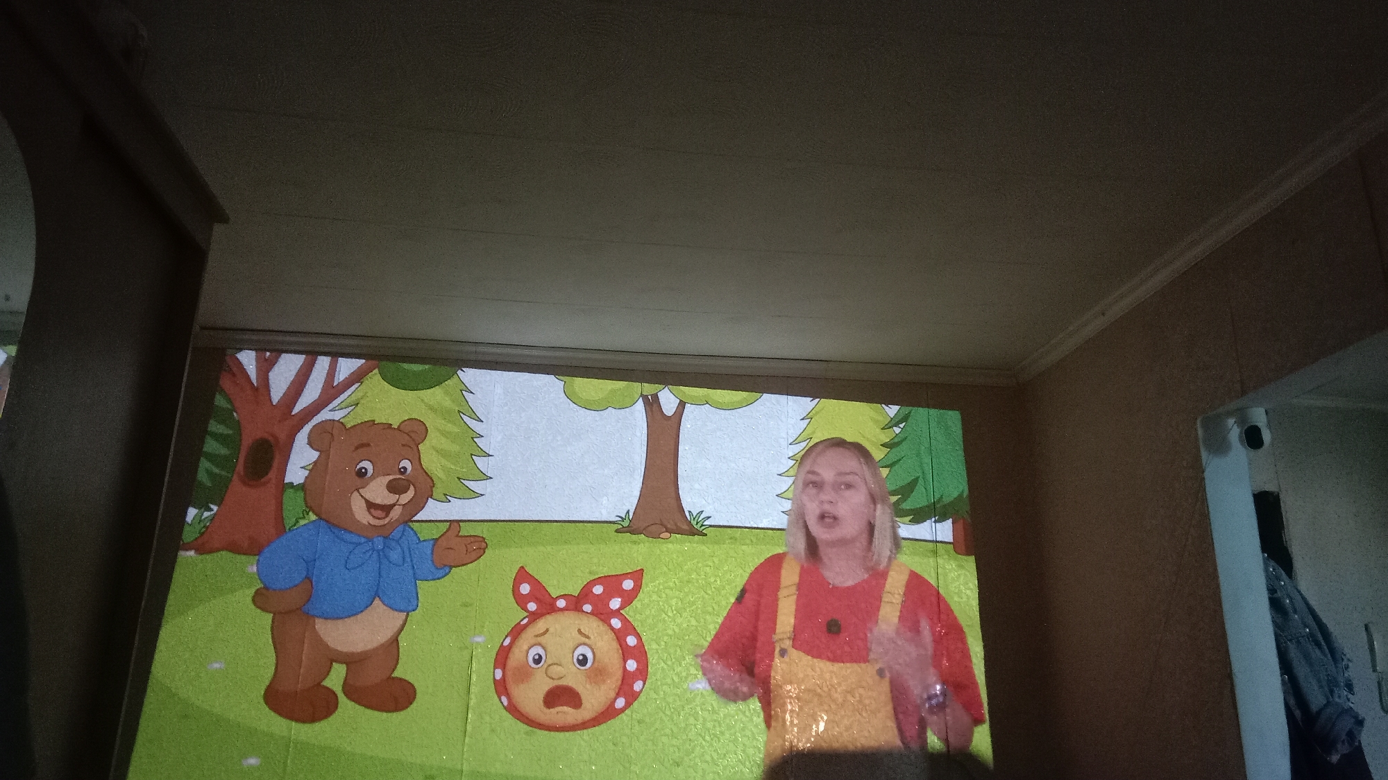 In Petropavl, Kazakhstan, a projected image displays a cartoon scene featuring a brown bear wearing a blue shirt and a distressed yellow character with a red bow. Beside the projection, a young woman with blonde hair, wearing a red shirt and yellow overalls, stands and gestures. The projection is cast onto a wall, which appears to be a projection screen, within a room that has textured walls and a textured ceiling. The overall lighting conditions suggest an indoor environment, possibly a home or classroom, with the projection being the primary light source in the immediate area.