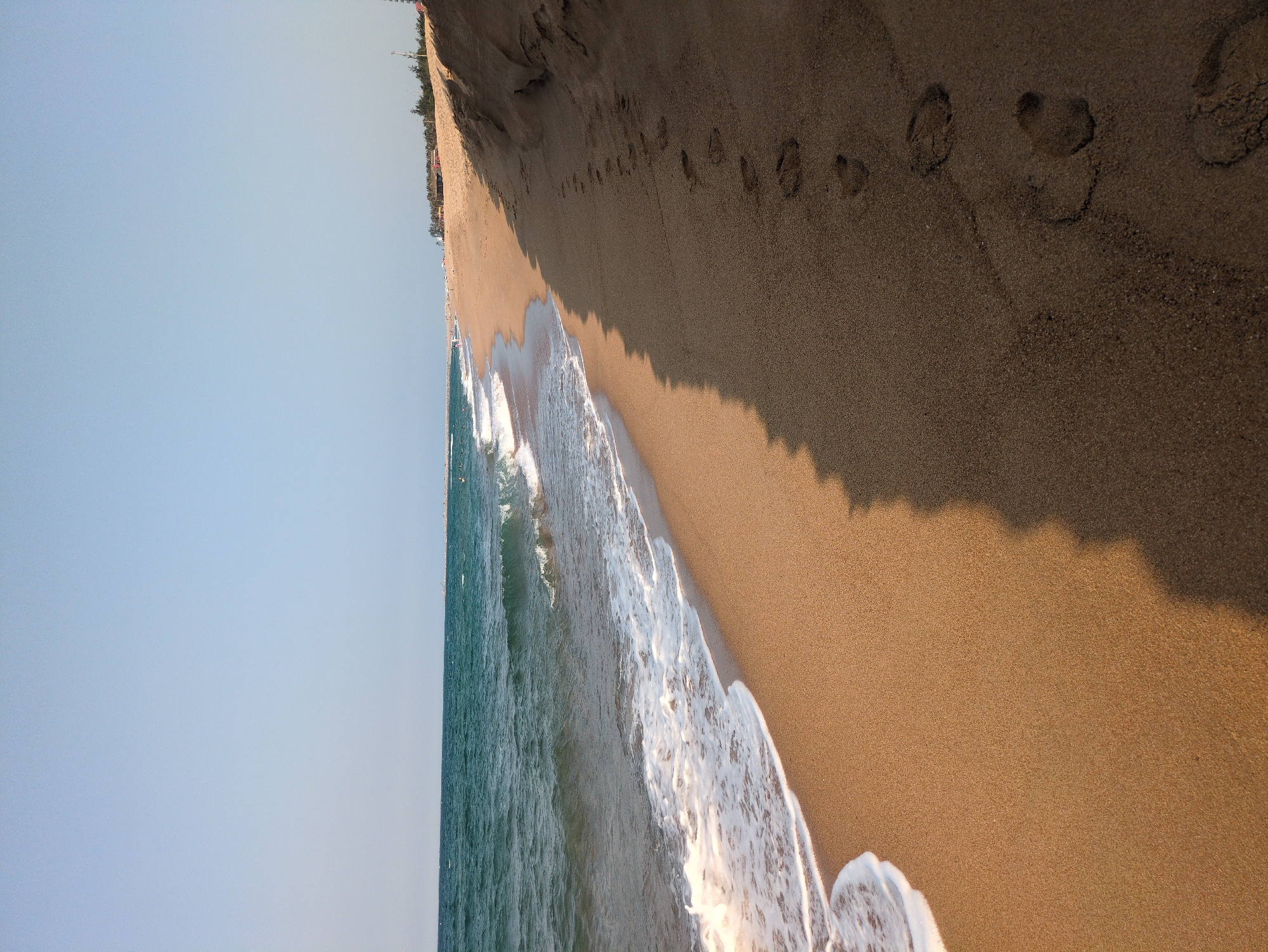 The image depicts a beach scene in Tuy Hòa, Vietnam. The scene is divided between the blue sky and ocean on the left and the sandy beach on the right. The ocean waves are crashing onto the shoreline, creating a foamy white border against the tan sand. Footprints are visible in the sand, indicating recent activity. The upper part of the beach transitions into a darker, coarser sand, likely leading to dunes. In the far distance, indistinct structures and vegetation suggest the edge of the city.