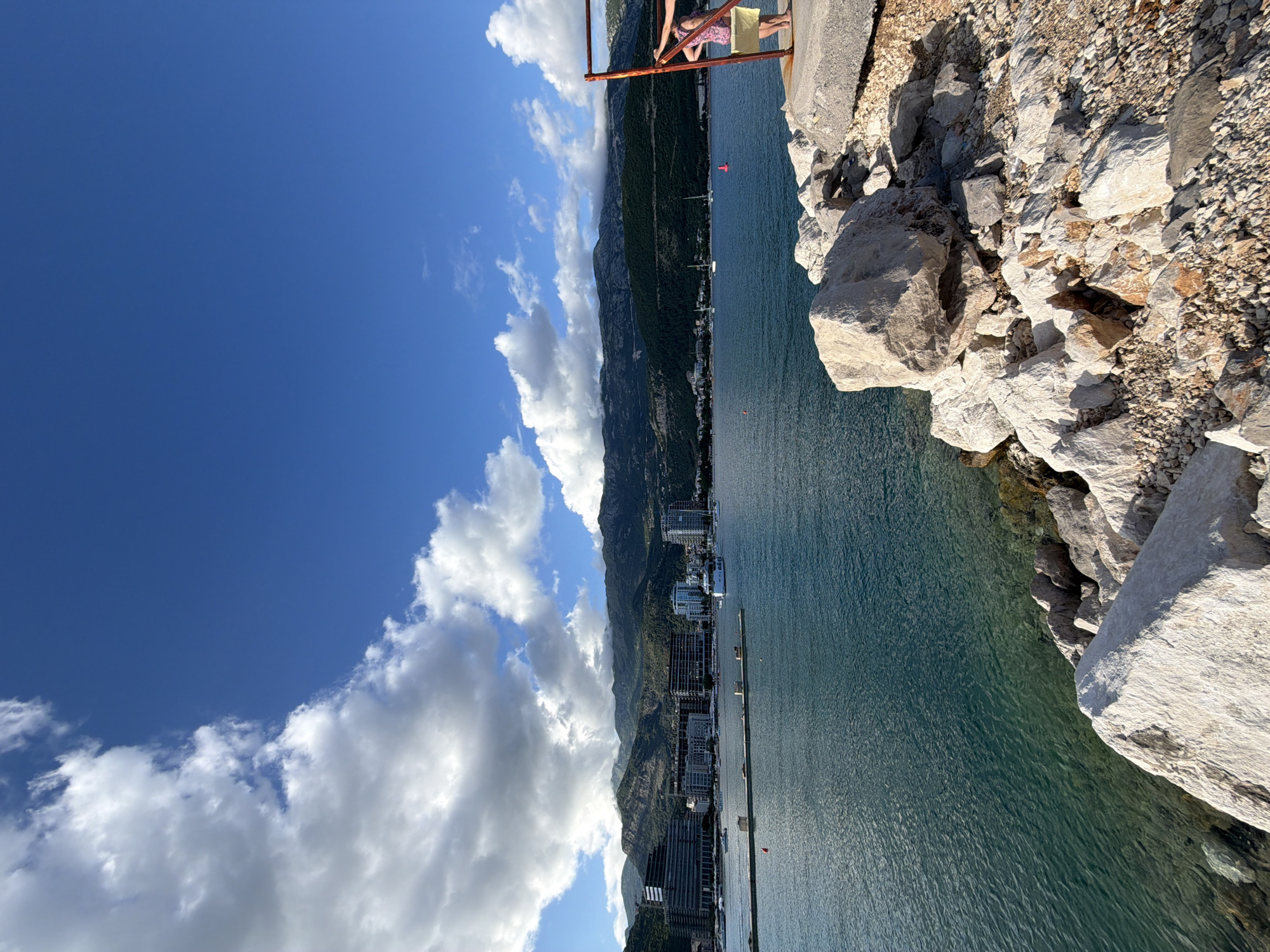 An outdoor photograph captures a scenic coastal landscape in Budva, Montenegro, under bright daylight. The image is taken from an elevated perspective, looking out over a bay or sea inlet.
In the foreground, on the right side, there is a rocky, uneven terrain with large light-colored boulders and smaller gravel. A rusty, reddish-brown metal barrier or railing is visible in the upper right. Partially visible behind this railing is one person, likely female, whose legs and lower torso are seen. They appear to be wearing a light-colored, possibly patterned top and light shorts or a skirt, standing and observing the view.
The middle ground features a calm, deep blue-green body of water, likely the Adriatic Sea. Several small boats, appearing as white specks, are scattered across the water. A few red buoys are also visible. Along the distant coastline, a town with numerous modern buildings, some appearing as multi-story structures, stretches out.
In the background, majestic dark mountains rise steeply from the coastline, creating a dramatic backdrop to the town and water. The sky above is a vibrant clear blue, dotted with scattered white, fluffy clouds. The overall impression is one of a sunny day with good visibility. There is no visible text in the image.