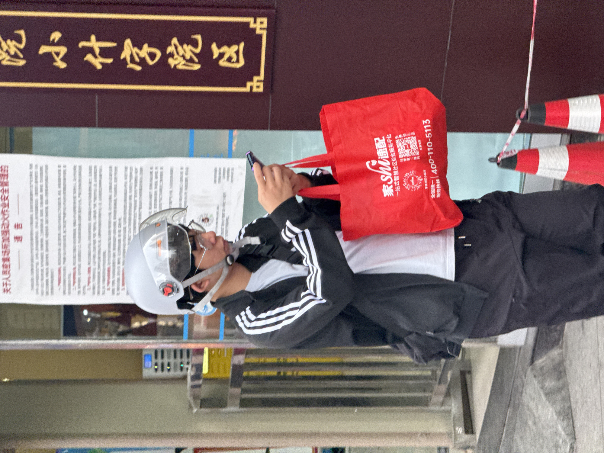 A man stands outside a building in Jiangjin, China. He wears a helmet, glasses, and a jacket with three white stripes on the sleeves, holding a cell phone in one hand and a red tote bag in the other. The tote bag has the Chinese characters "家Shi速配" and a service hotline printed on it. Behind him, a sign is posted on the building with Chinese characters. Red and white cones are partially visible in the background.