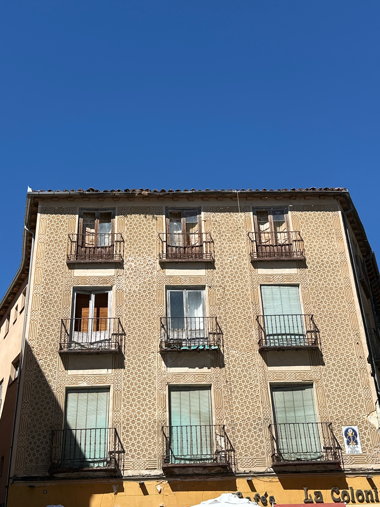 The image shows the facade of a building in the city of La Lastrilla, Spain. The building has a patterned facade with a light beige base color and a decorative pattern in a slightly darker shade. There are nine windows visible on the facade, arranged in three rows of three. Each window has a small balcony with a wrought-iron railing. Some of the windows are covered with blinds, while others are open, revealing wooden shutters or curtains. The roof is tiled, and the sky above is a clear blue. To the right of the building, a small blue and white tile with a crest and text is visible. Below the building, a yellow storefront with red lettering that reads "La Coloni" is partially visible. The overall scene suggests an architectural detail within a European urban environment.