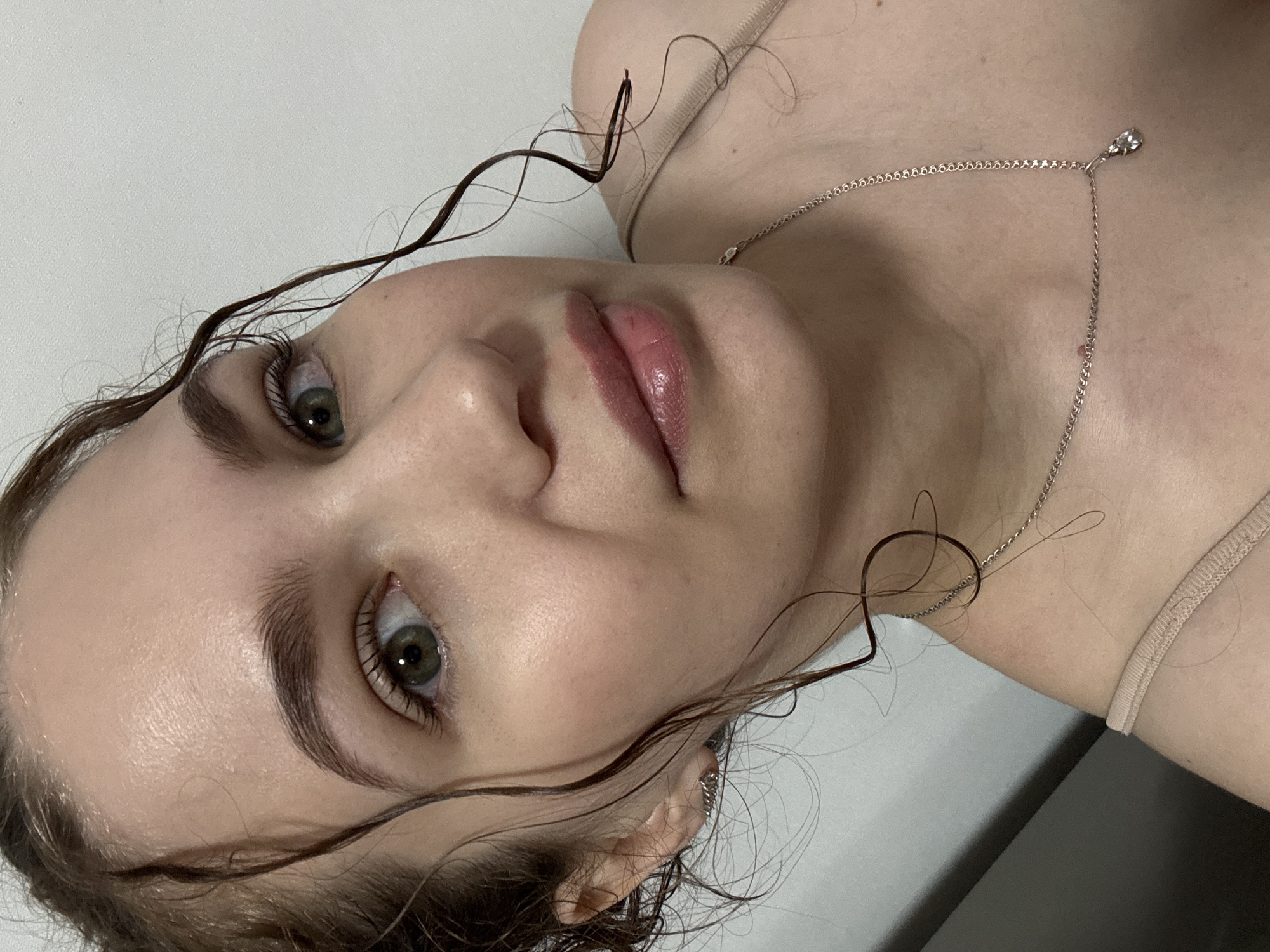 A close-up portrait shows a young woman from an overhead, rotated perspective, with her head resting on a plain, light-colored surface, possibly a bed or floor, looking towards the camera. Her face dominates the frame, showcasing her fair complexion and prominent features. She has striking green or hazel eyes, enhanced by dark eyelashes with visible mascara, and well-defined, dark eyebrows. Her lips are full, glossy, and tinted with a reddish-pink color.
Her dark brown, wavy hair, some strands appearing damp or artfully styled, frames her face and curls around her ear. A delicate silver-toned necklace with a small, clear gemstone pendant adorns her décolletage, resting just above the visible strap of a light beige or nude-colored top or undergarment. The lighting is soft and even, highlighting her smooth skin which has a subtle sheen. The background is an unadorned light surface, providing no specific location cues other than an indoor setting. There is no visible text. The overall mood is calm and introspective, capturing a moment of relaxed posing. This scene is located in Chaikovsky, Russia.