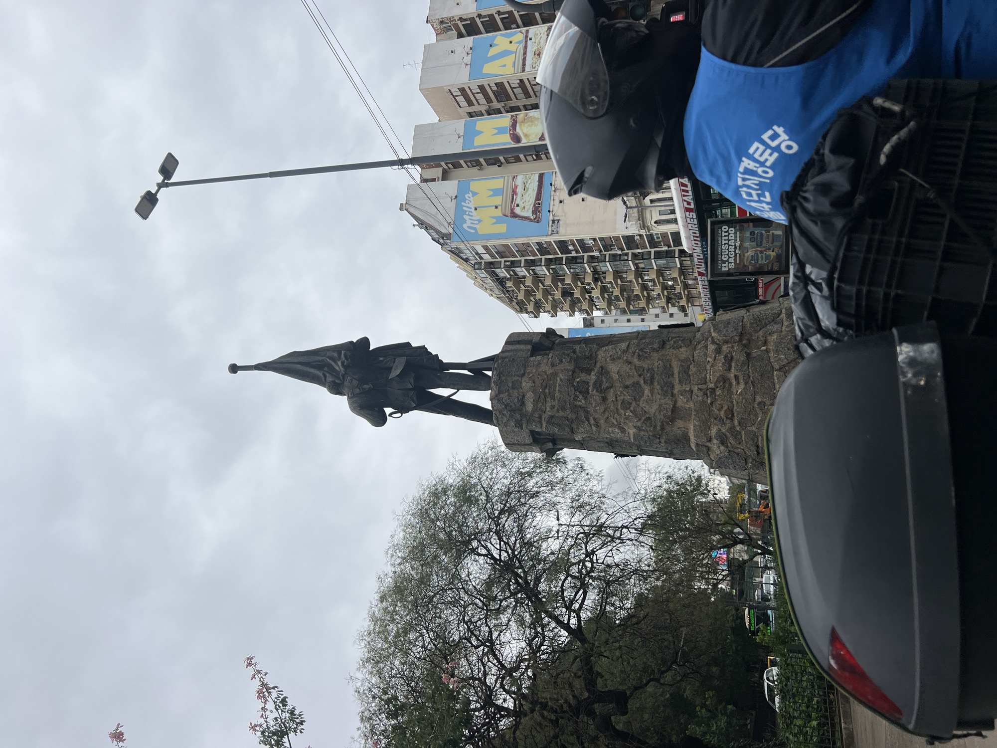 The image depicts a view of the monument to Christopher Columbus in Buenos Aires, Argentina. The bronze statue of Columbus, holding a flag, stands atop a stone pedestal. Behind the statue, a building with a large "Milka" advertisement is visible. A street lamp is positioned above the statue. In the foreground, partially obscuring the view, is a person on a motorcycle wearing a helmet and a blue vest with Korean writing. A motorcycle storage box is also visible. The sky is overcast. Trees are visible at the base of the monument. Buses are visible in the background.
