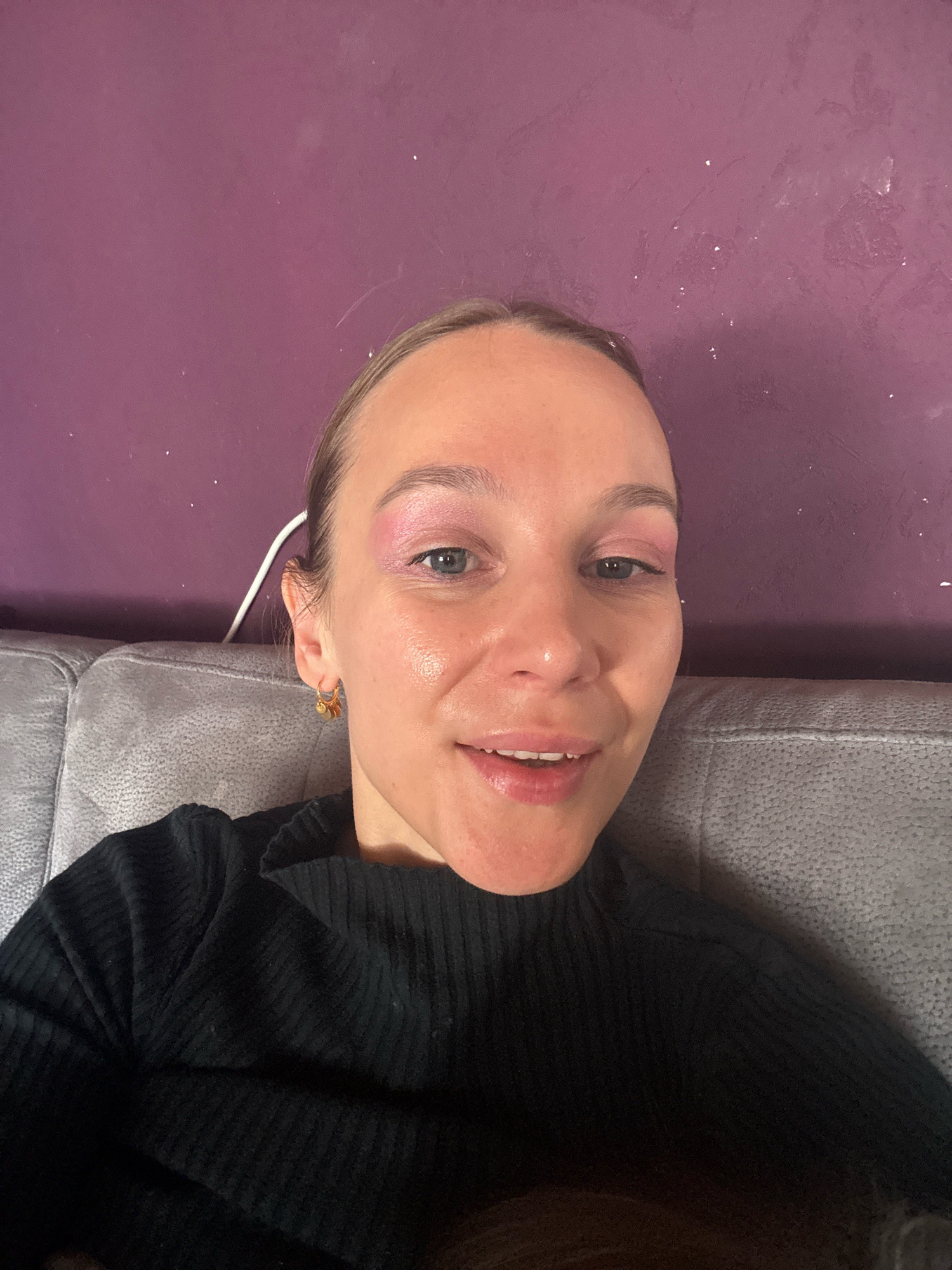 A young woman with blonde hair pulled back and wearing a black ribbed top is seated on a grey sofa in front of a textured purple wall. She is looking at the camera and smiling with her mouth slightly open, revealing her teeth. Her makeup includes pink eyeshadow and lipstick. She is wearing a small gold hoop earring with red accents. A white cord is visible behind her ear. The scene is likely indoors, possibly in a residential setting in Überherrn, Germany.