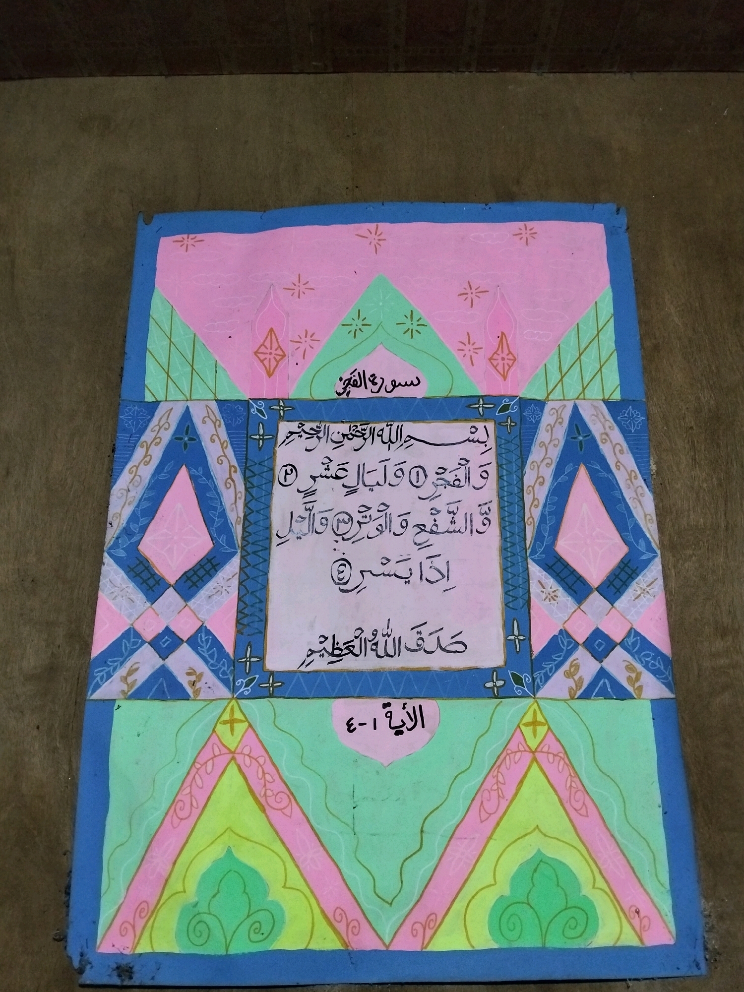 The image shows a colorful, hand-painted canvas featuring Arabic calligraphy. The calligraphy is centered on a light pink background with the words "بِسْمِ اللهِ الرَّحْمٰنِ الرَّحِيْمِ" (In the name of Allah, the Most Gracious, the Most Merciful) at the top. Below this are verses of the Quran, indicated by numbers within circles: "وَالْفَجْرِ" (and the dawn), "وَلَيَالٍ عَشْرٍ" (and the ten nights), "وَالشَّفْعِ وَالْوَتْرِ" (and the even and the odd), and "وَالَّيْلِ إِذَا يَسْرِ" (and by the night when it passes). At the bottom of the calligraphy section, it says "صَدَقَ اللهُ الْعَظِيمُ" (Allah the Almighty has spoken the truth). The canvas is framed by geometric patterns in pink, blue, and green. The bottom section displays the text "الْآيَةِ ٤-١" (Verse 4-1). The canvas is laid flat on a textured, brown surface, likely a wooden floor or ground. This object appears to be a piece of religious art, possibly a decorative mat or a section of a larger display. The specific location in Teluk Pambang, Indonesia, is not evident from the image itself.