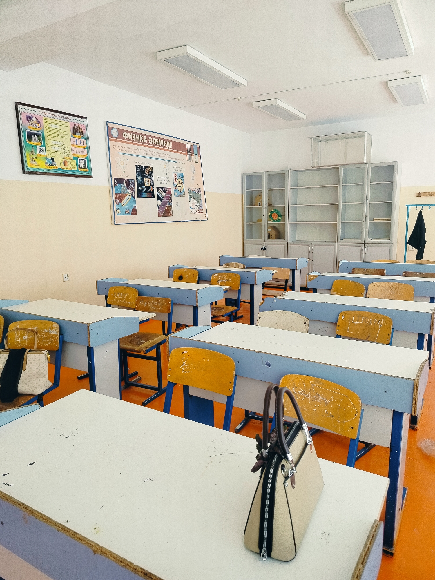 The image depicts an empty classroom in Көлкент, Kazakhstan. The room contains rows of desks with blue trim and yellow seats. Whiteboards are mounted on the walls, along with educational posters. To the left, a poster titled "ГЕОМЕТРИЯЛЫҚ ОПТИКА" is visible, and to the right, a poster with the title "ФИЗИКА ӘЛЕМІНДЕ" is displayed. Tall, glass-fronted cabinets are situated against the back wall, filled with shelves. A black jacket hangs on a hook on the right side of the back wall. The floor is a bright orange color, and fluorescent light fixtures are installed on the ceiling. In the foreground, a beige handbag with brown handles rests on a desk. There are no people visible in the image.