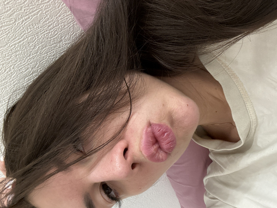 The image displays a close-up, angled portrait of a person, seemingly a selfie, taken from an overhead perspective while the person is lying down or tilting their head. The subject's head is angled downwards towards the bottom left of the frame.
The most prominent feature is the person's face, specifically their lips, which are pursed into a distinct "kissing" or "duck face" expression. The lips are full, a natural pink color, and show visible texture and vertical lines. Above the lips, the tip and bridge of a nose are visible. One eye, the left one, is partially visible towards the bottom left of the frame, looking towards the viewer, with visible dark eyelashes. The person's skin appears smooth and has a light complexion.
Long, dark brown hair cascades across the left side of the face and over the shoulder. Some strands fall over the forehead and near the visible eye. On the right side of the frame, a portion of a light-colored, possibly off-white or cream-colored, top or t-shirt with a ribbed collar is visible, covering the person's shoulder and neck area.
The background consists of two distinct areas. To the left of the hair and face, there is a textured white wall, possibly wallpaper. Behind the head and extending to the top right, a soft, plain light purple surface, likely a pillow or bedding, is visible. The overall lighting is soft and even, suggesting an indoor setting. The image's tilted orientation creates a dynamic and casual feel.