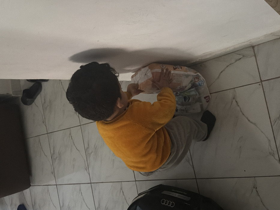 An indoor scene in Dar Bouazza, Morocco, depicts a young child from a high-angle perspective. The child, wearing a mustard-yellow long-sleeved top and light grey sweatpants, is in a crouched position, facing towards a white interior wall. Their dark, curly hair is visible. Both hands are placed on a large, clear plastic-wrapped package of disposable diapers, which is resting against the wall. The diaper package displays the number "38" and other partially legible branding. The floor is composed of light-colored square tiles with dark, irregular veining patterns. In the upper left background, a black object with a silver "AUDI" logo is visible, alongside a dark slipper and a piece of brown furniture. Shadows are cast on the white wall to the child's right.