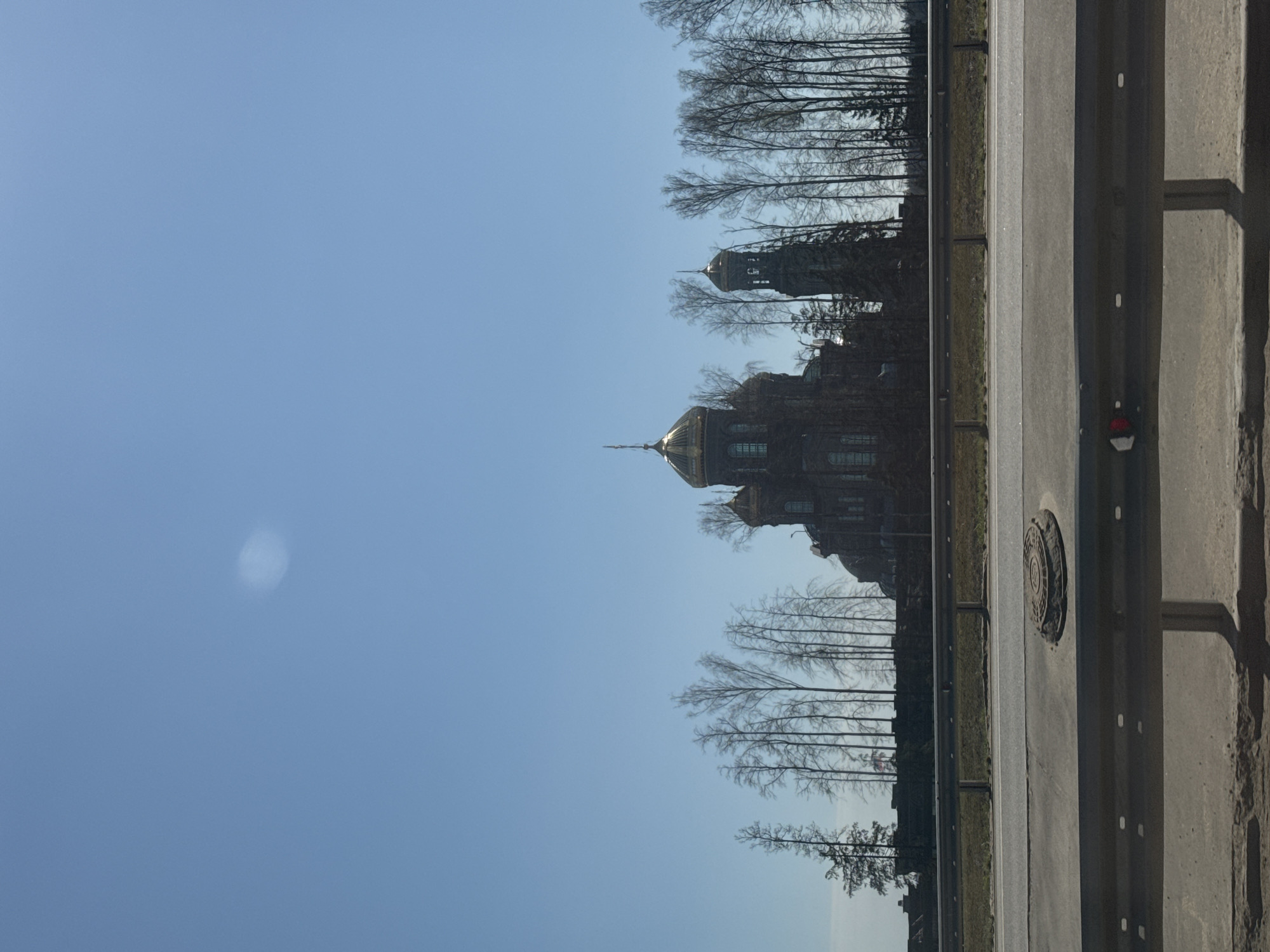 The image presents a view with the sky occupying the majority of the space. An ornate church with golden domes and a bell tower can be seen, partially obscured by bare trees, likely indicating the season is late autumn or winter. To the right of the church and trees is a highway, complete with guardrails and road markings. There is also a manhole cover visible on the edge of the highway, which may suggest that the scene is near a urban area, but the exact location is not evident from the image.