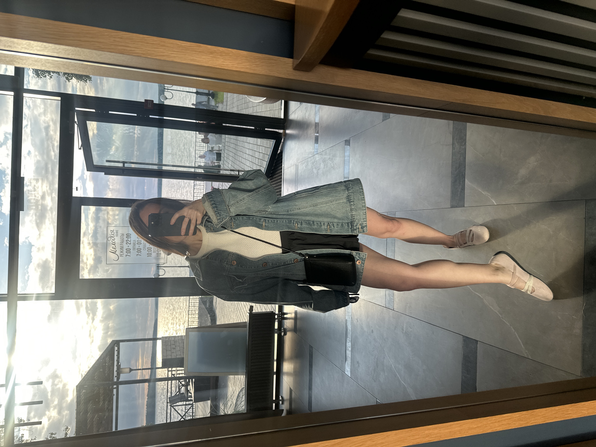 The image shows a woman taking a mirror selfie in what appears to be a restaurant or cafe with large windows overlooking a body of water. The woman is wearing a denim jacket, a white top, a black skirt, and light-colored flat shoes. She is holding a black smartphone to take the picture. A small black purse hangs from her shoulder.
Through the windows, the scene outside suggests it is late afternoon or early evening, with the sun casting a warm glow and creating reflections on the water. The sky is partly cloudy. In the reflection on the window, we can see people walking outside and some outdoor seating areas.
Visible on the window is a sign with Russian text that appears to be the operating hours for a place called "Мелодия" (Melody). The hours listed are 7:00-22:00, with extended hours on Friday (7:00-00:00) and Saturday (10:00-). The reflection of the interior shows a tiled floor and wooden architectural elements. The overall impression is a casual, stylish setting during a pleasant time of day.