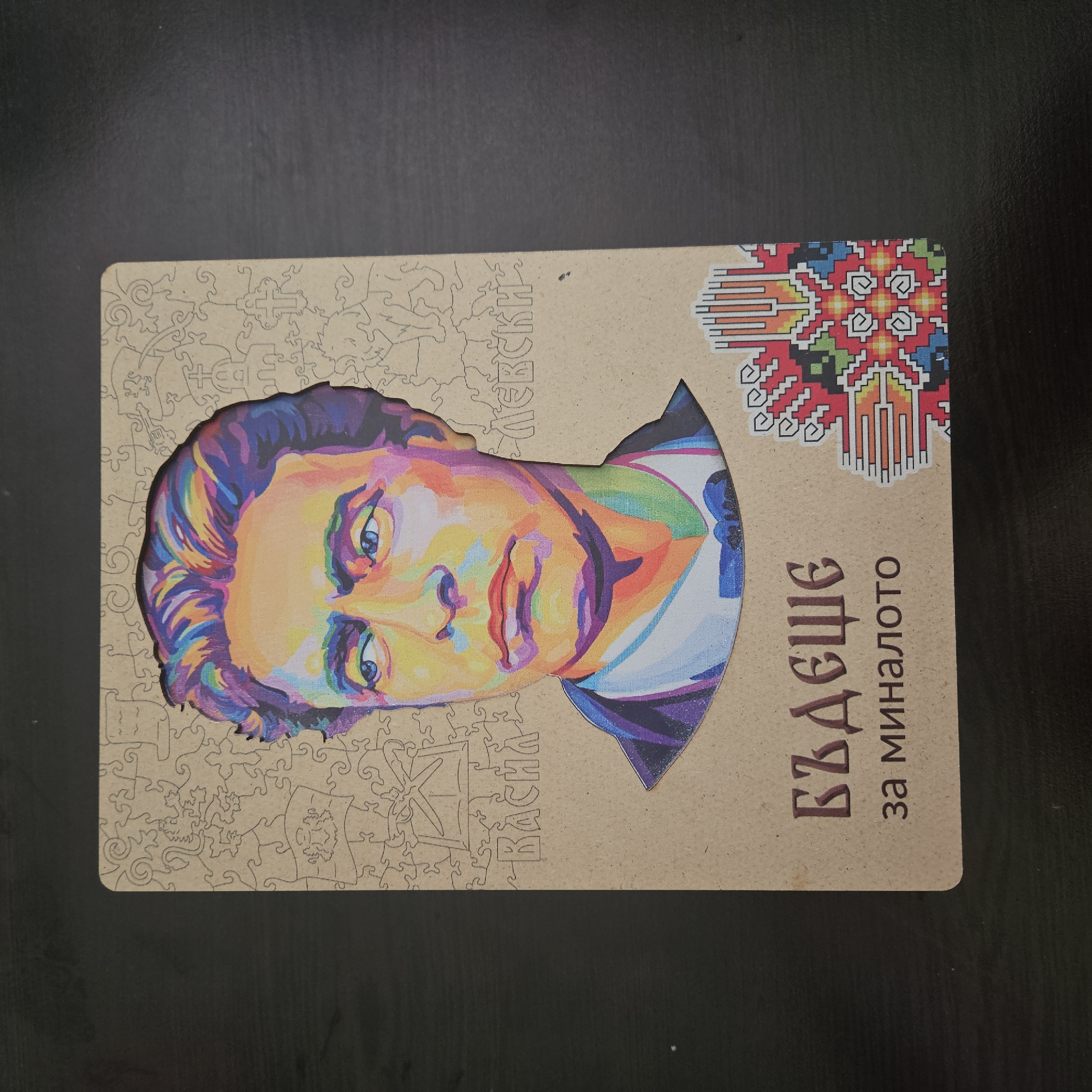 The image shows a portrait of Vasil Levski, a Bulgarian revolutionary and national hero. The portrait is stylized with bright, non-realistic colors and is surrounded by a jigsaw puzzle-like border. The background is a plain, likely wooden surface, which appears to be part of the design or the base upon which the portrait rests. The bottom right corner contains Bulgarian Cyrillic text. To the right of the portrait's head is a traditional Bulgarian embroidery design. The city of Stara Zagora, Bulgaria, is relevant because Levski established a revolutionary committee there.