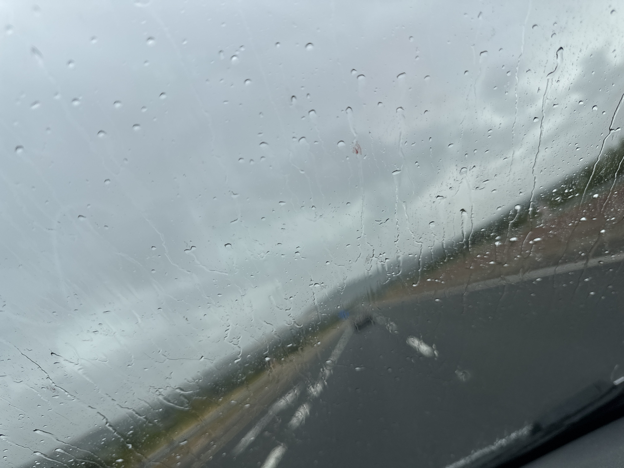 The image, taken from inside a vehicle, shows a view through a rain-streaked windshield on a cloudy day. The road ahead is visible, with lane markings and what appears to be the side of the road with some greenery. Water droplets of various sizes are scattered across the glass, distorting the view and creating streaks as they run down. In the distance, a small red object is visible on the windshield, possibly a sticker or debris. The sky is overcast, suggesting a possibility of rain or recent rainfall. The scene appears to be on a highway or a similar road, with a sense of motion implied by the blurred background. No specific landmarks or details indicate the location within Unknown, Ireland.