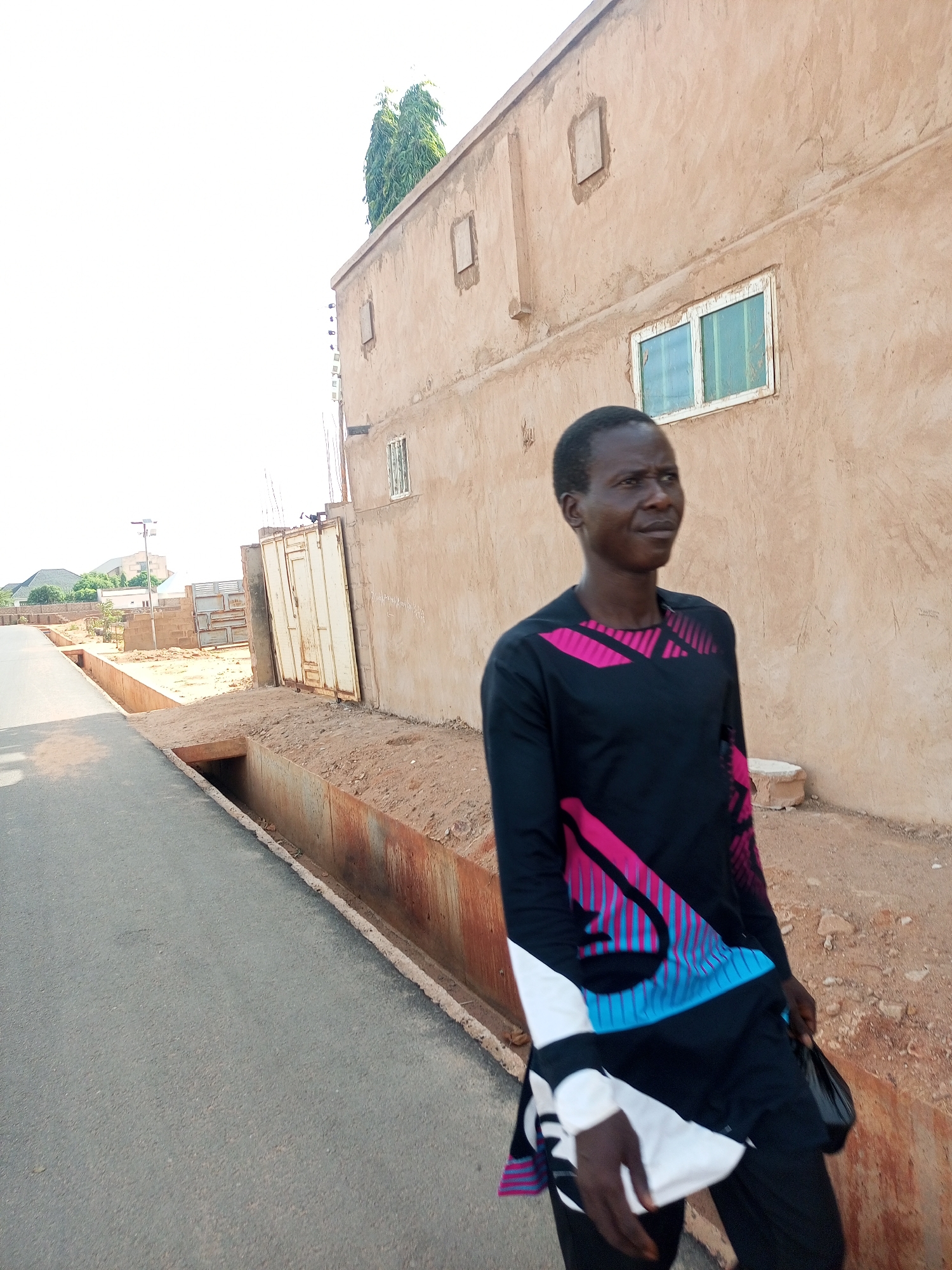 A man stands on the side of a paved road in Gombe, Nigeria. He is wearing a black long-sleeved shirt with pink and blue graphic designs, and black pants. Beside him is a drainage ditch, and behind him is a building with small, framed windows. In the background, another building is visible, and some sparse vegetation is present. The sky is bright, indicating a daytime scene.