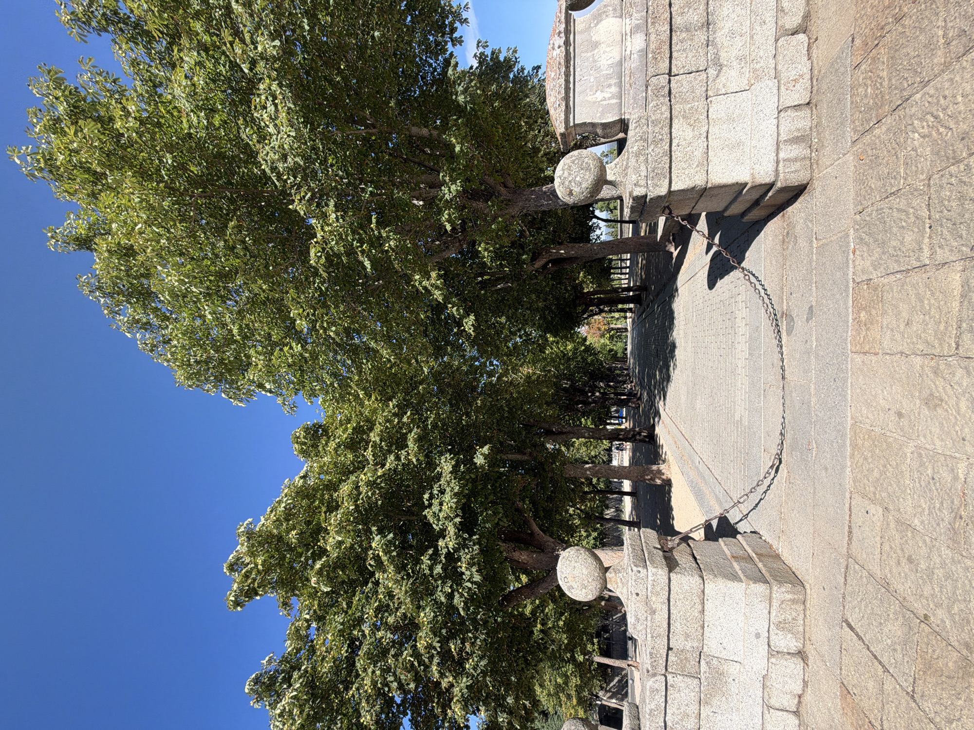 The image depicts an outdoor scene in the city of San Lorenzo de El Escorial, Spain, on a clear day with a bright blue sky. The foreground is dominated by a paved walkway constructed of large, irregular stone slabs. This walkway leads into a tree-lined avenue. Flanking the entrance to this avenue are two substantial stone pillars. These pillars are made of stacked, roughly cut rectangular stone blocks, topped with spherical finials carved from stone. Connecting these two pillars is a heavy metal chain, draped between them and resting on the stone paving.

The tree-lined avenue features a path of similar stone paving, bordered by sand or gravel. Mature trees with dense green foliage line both sides of this path, creating a canopy overhead. The sunlight casts shadows from the trees onto the path, indicating a sunny day. In the distance, along the avenue, a few individuals can be seen walking. The overall impression is one of a well-maintained public space, likely a park or promenade, within the city. The architecture of the stone pillars and the paving suggest historical significance.