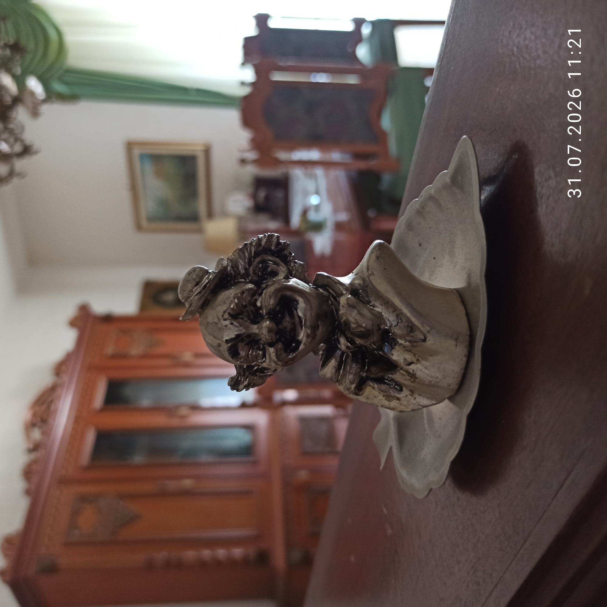 A vintage, metallic clown figurine with exaggerated features and a wide smile sits on a decorative, scalloped plate. The figurine is positioned on a polished wooden surface, likely a table or dresser. In the background, a large, ornate wooden cabinet with glass doors is visible, suggesting an indoor setting such as a home or antique shop. The overall ambiance is one of a dimly lit room, possibly an older home, with natural light filtering in from a window covered by green drapes on the right side of the frame. A timestamp "31.07.2026 11:21" is present in the lower right corner of the image. The location is identified as Modugno, Italy.