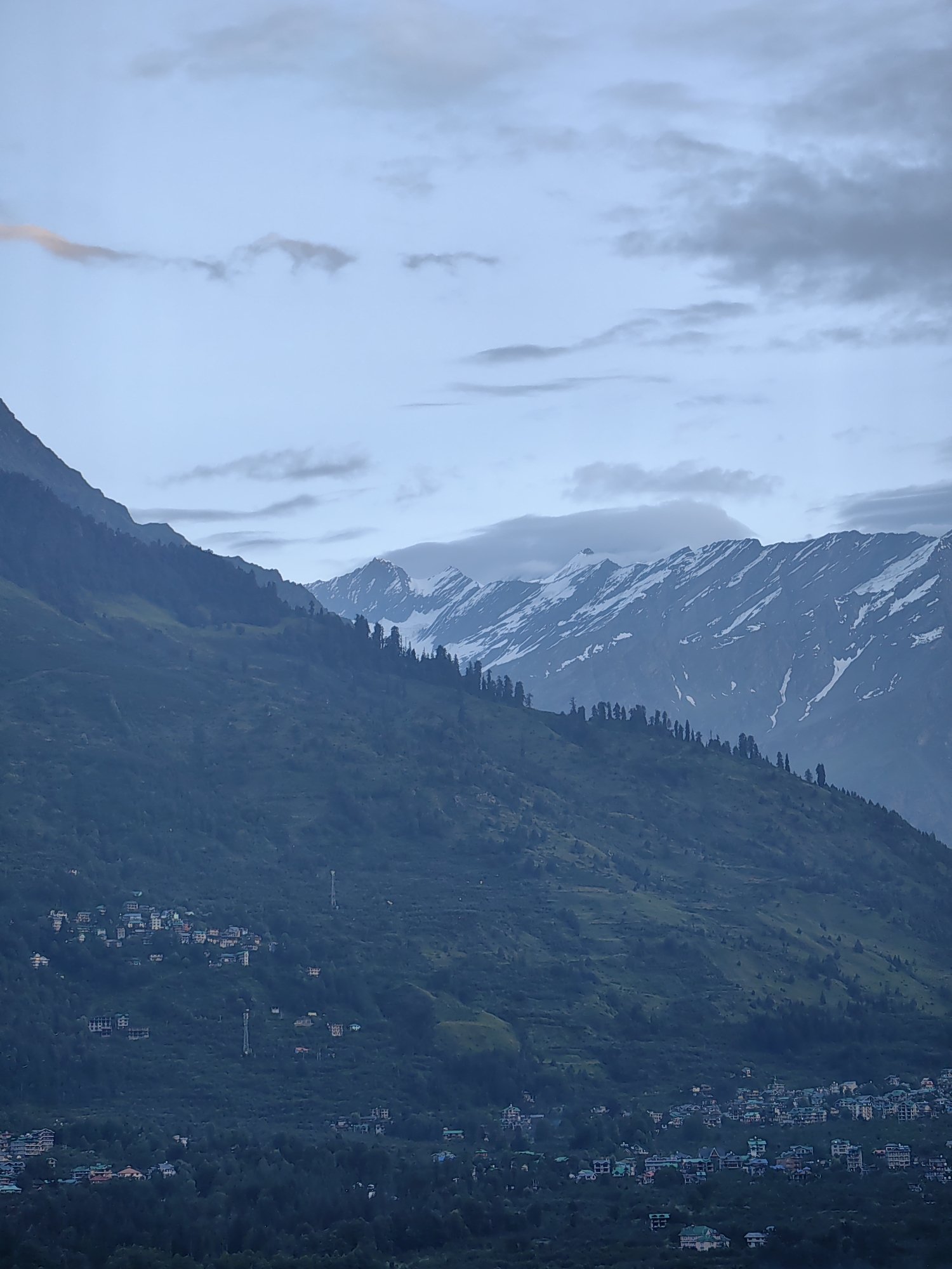 The image shows a panoramic view of the town of Vashisht, India, nestled amidst the Himalayan mountains. The foreground is dominated by a lush green mountainside, dotted with scattered trees and the rooftops of houses. As the eye moves upwards, the landscape transitions to higher altitudes, revealing a majestic mountain range. The peaks are capped with snow, their rugged slopes streaked with white. The sky above is overcast, with soft, diffused light indicating either early morning or late evening. The town of Vashisht is visible in the mid-ground, with clusters of buildings spread across the lower slopes of the mountains. The architecture appears to be a mix of traditional and modern styles, with colorful roofs standing out against the muted tones of the natural environment. There are no people or animals visible in the image, and no specific actions or interactions are depicted. The overall scene conveys a sense of tranquility and the grandeur of nature.