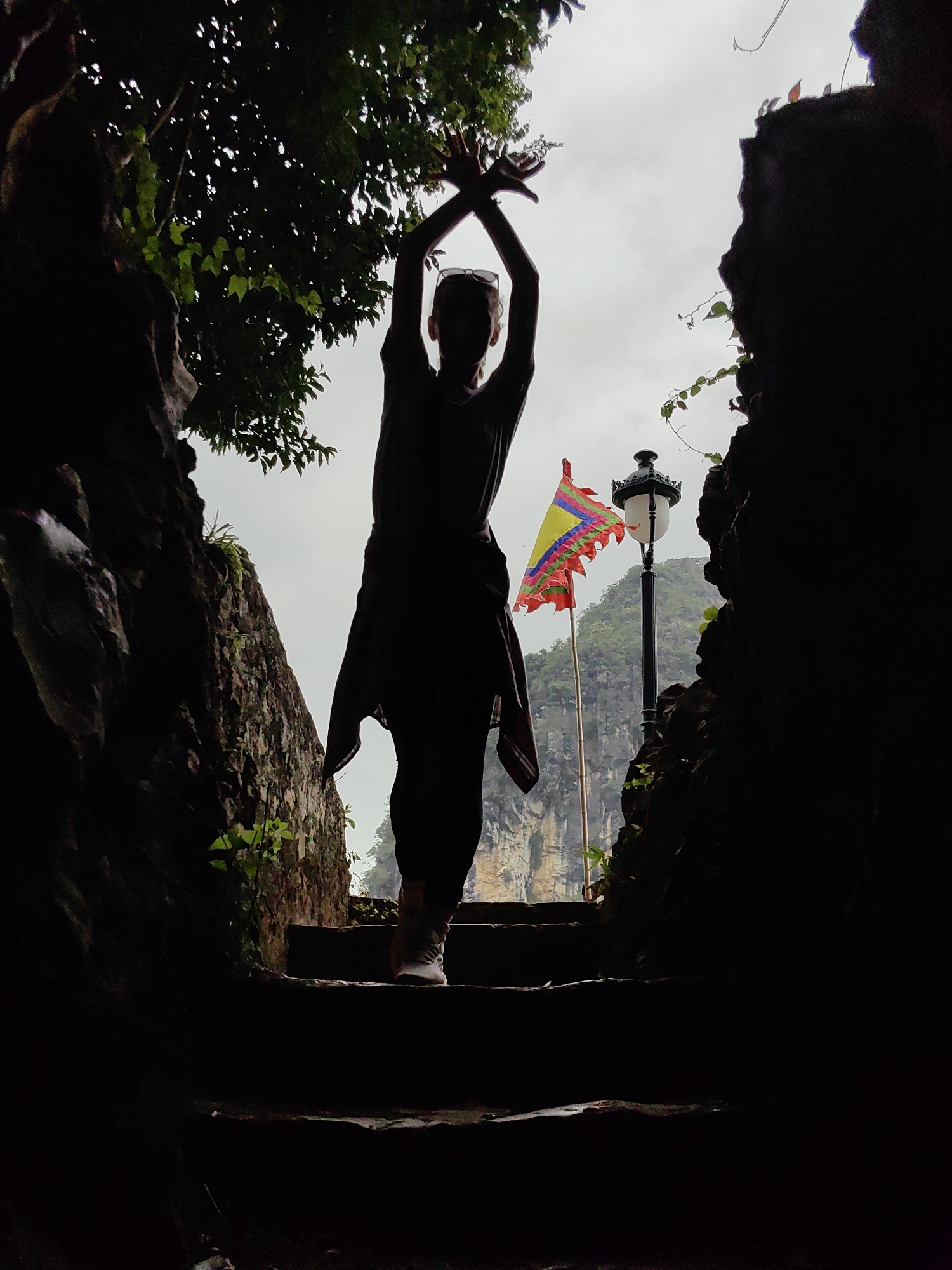 A silhouetted individual stands in a stone passageway, their arms raised and crossed above their head. The passageway leads to an opening revealing a colorful flag, a lamppost, and a mountain in the distance. The scene is observed on cloudy day. The location is in Lạng Sơn, Vietnam.