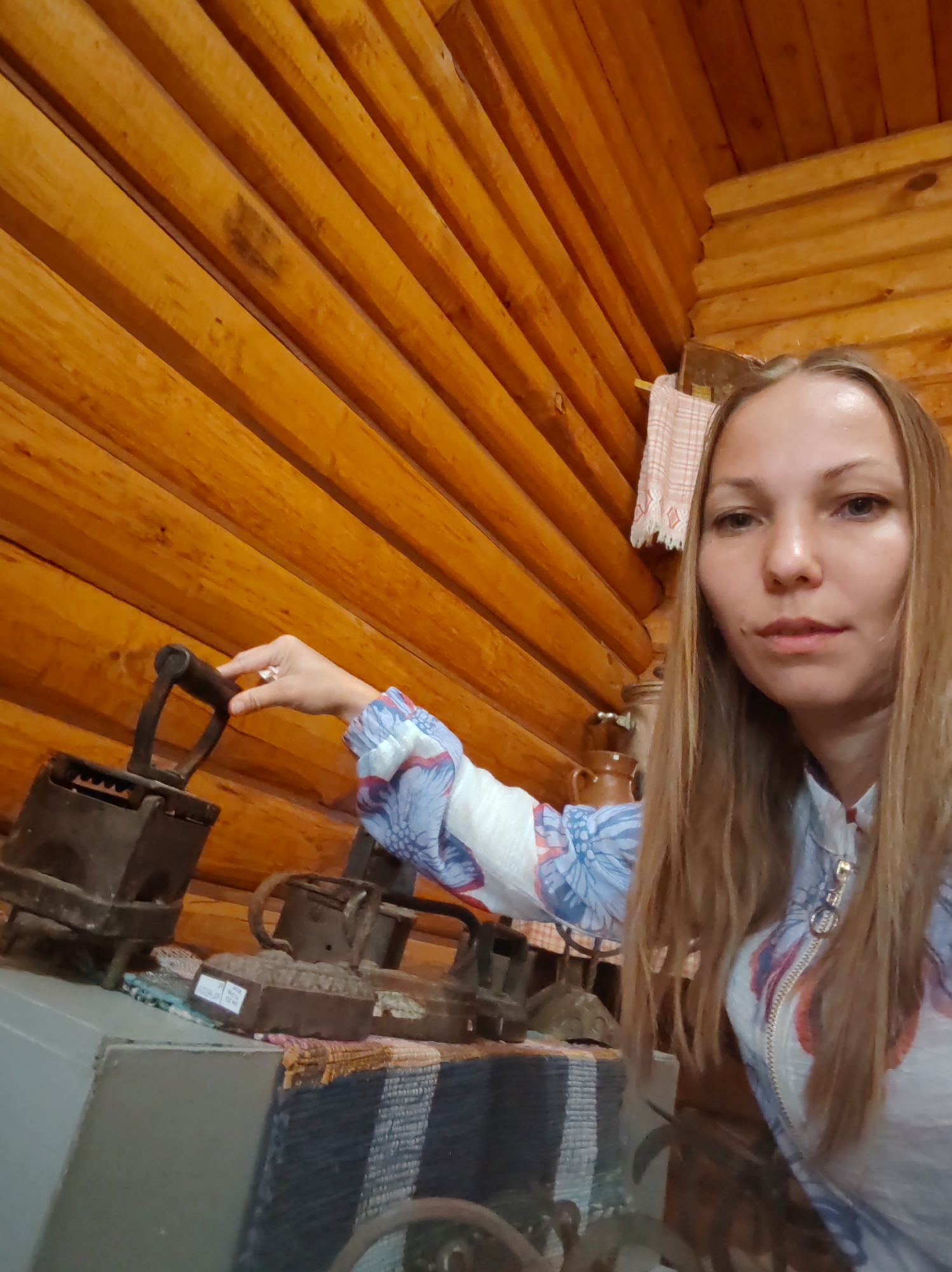 A long shot, taken from eye level, shows a woman with long, blonde hair posing for a selfie in a room with wooden walls. She is wearing a white blouse with a blue and red floral pattern and a zipper. Her right hand is resting on the handle of an antique iron. In the background, there are several more irons, a jug, and a towel hanging on the wall. The room appears to be a museum or historical exhibit, given the display of old irons. The wooden walls are made of horizontal logs, a common feature in rural Russian architecture. The lighting is natural and soft, coming from the right side of the frame. The woman's expression is neutral, and she is looking directly at the camera. The overall impression is of a casual snapshot taken during a visit to a local attraction in Vizinga, Russia.