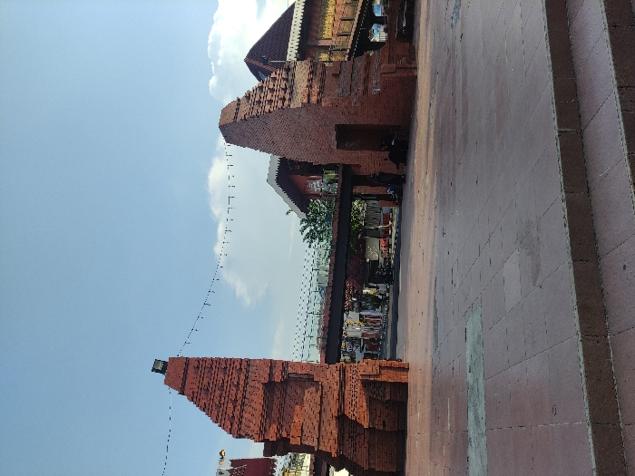 The image depicts a red brick structure, possibly a remnant of a historic building, located in Mojokerto, Indonesia, at coordinates -7.464 latitude and 112.432 longitude. The structure is composed of two tall, rectangular brick pillars, each with a distinct, slightly angled shape. A metal bridge, with a black railing, connects the two pillars, extending over a paved area. The bridge appears to be part of a pedestrian overpass. The structure is in a state of partial disrepair, with some of the bricks missing or cracked. There are several individuals, dressed in casual attire, standing or walking on the paved area beneath the bridge. The sky is a light blue, with white clouds scattered across it. The ground is composed of a mixture of grey concrete and red brick pavers. Green foliage is visible in the background. The scene is illuminated by natural daylight, suggesting a daytime setting.