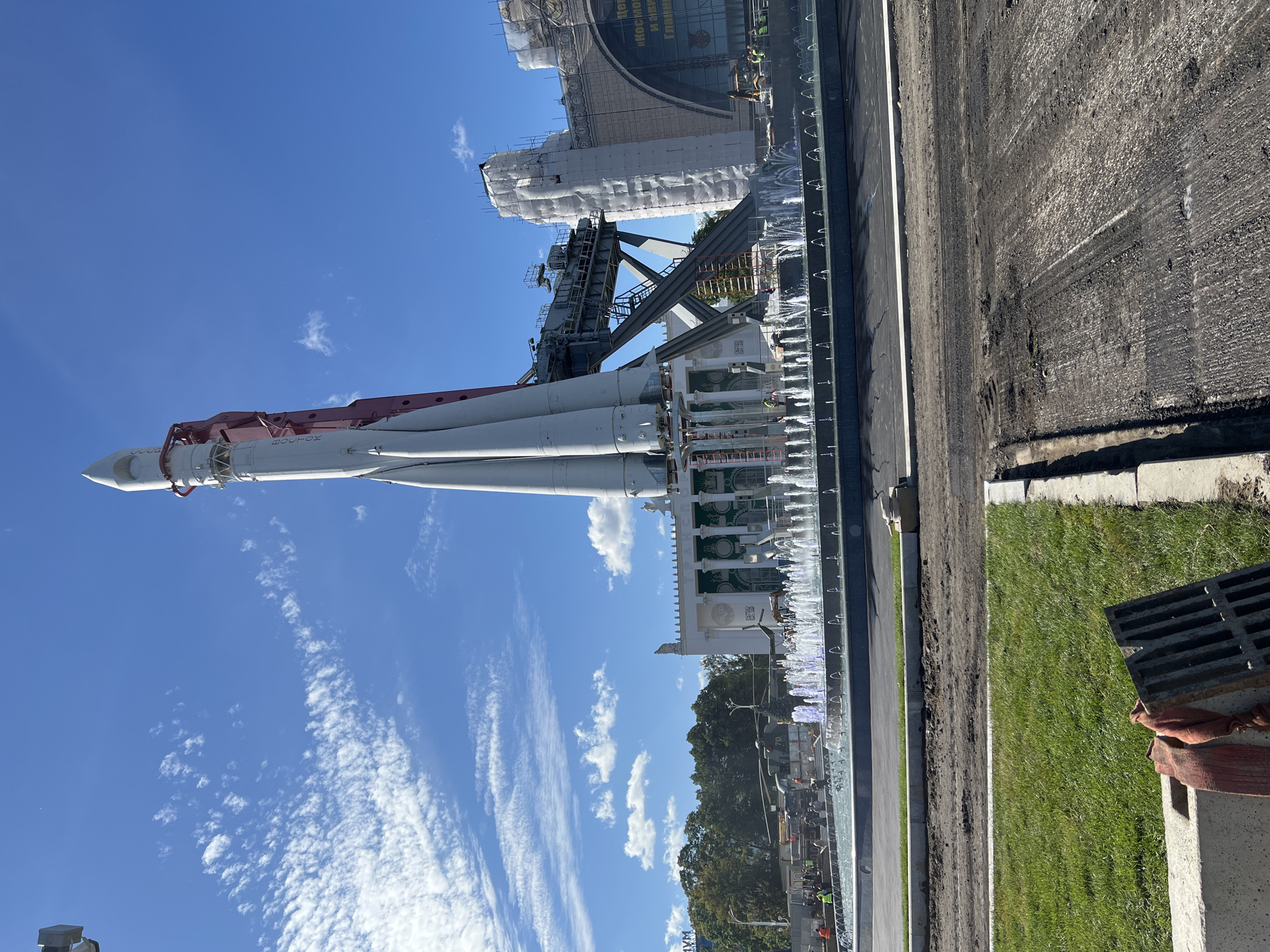 The image depicts the Vostok rocket monument located at VDNKh (Exhibition of Achievements of National Economy) in Moscow, Russia. The rocket, painted white and red, stands tall against a bright blue sky with scattered white clouds. It is positioned behind a large, multi-tiered fountain with numerous water jets spraying upwards. In the background, a grand pavilion with classical architectural elements and intricate detailing is visible. Portions of this pavilion appear to be under scaffolding and white sheeting, suggesting ongoing construction or renovation. Several trees and lush green grass areas surround the monument and pavilion. In the foreground, the ground is a mix of dark soil, possibly indicating recent landscaping or road work, and a patch of green grass with a metal grate and an orange cloth. Several people are scattered around the fountain and near the pavilion, some wearing yellow safety vests, suggesting they are workers. A helicopter and a truck are also visible in the background near the pavilion. The overall scene suggests a public space that is both a historical monument and an active area for maintenance and development.