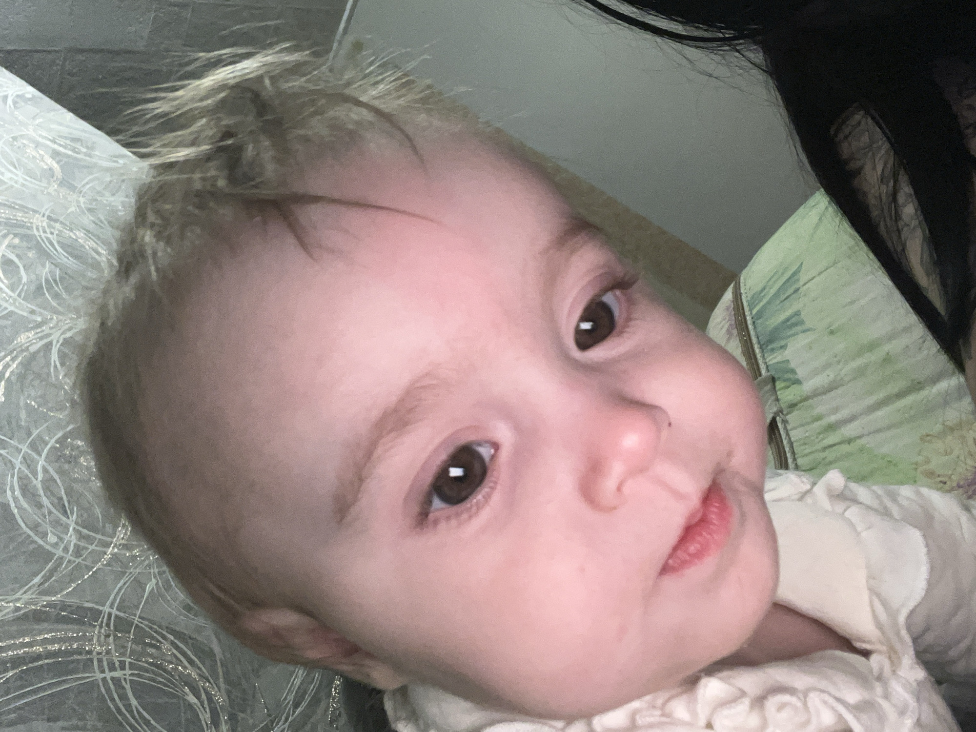 An infant with fair skin, brown eyes, and light brown hair is depicted in an indoor setting within Chaikovsky, Russia. The infant's face, positioned slightly off-center and tilted to the right, occupies the majority of the frame, looking directly towards the viewer. The subject wears a light-colored garment, possibly cream or off-white, with visible ruffles near the left shoulder. The infant's lips are slightly parted, revealing the lower lip. In the background, to the upper left, a wall covered in gray wallpaper with a silver, abstract swirling pattern is visible. Below the infant's neck and right shoulder, a green fabric with a subtle pattern is present, likely a blanket or pillow. A portion of dark hair, possibly belonging to an individual holding or accompanying the infant, is visible in the lower right corner of the frame. A plain light gray wall or surface is present in the upper right background.