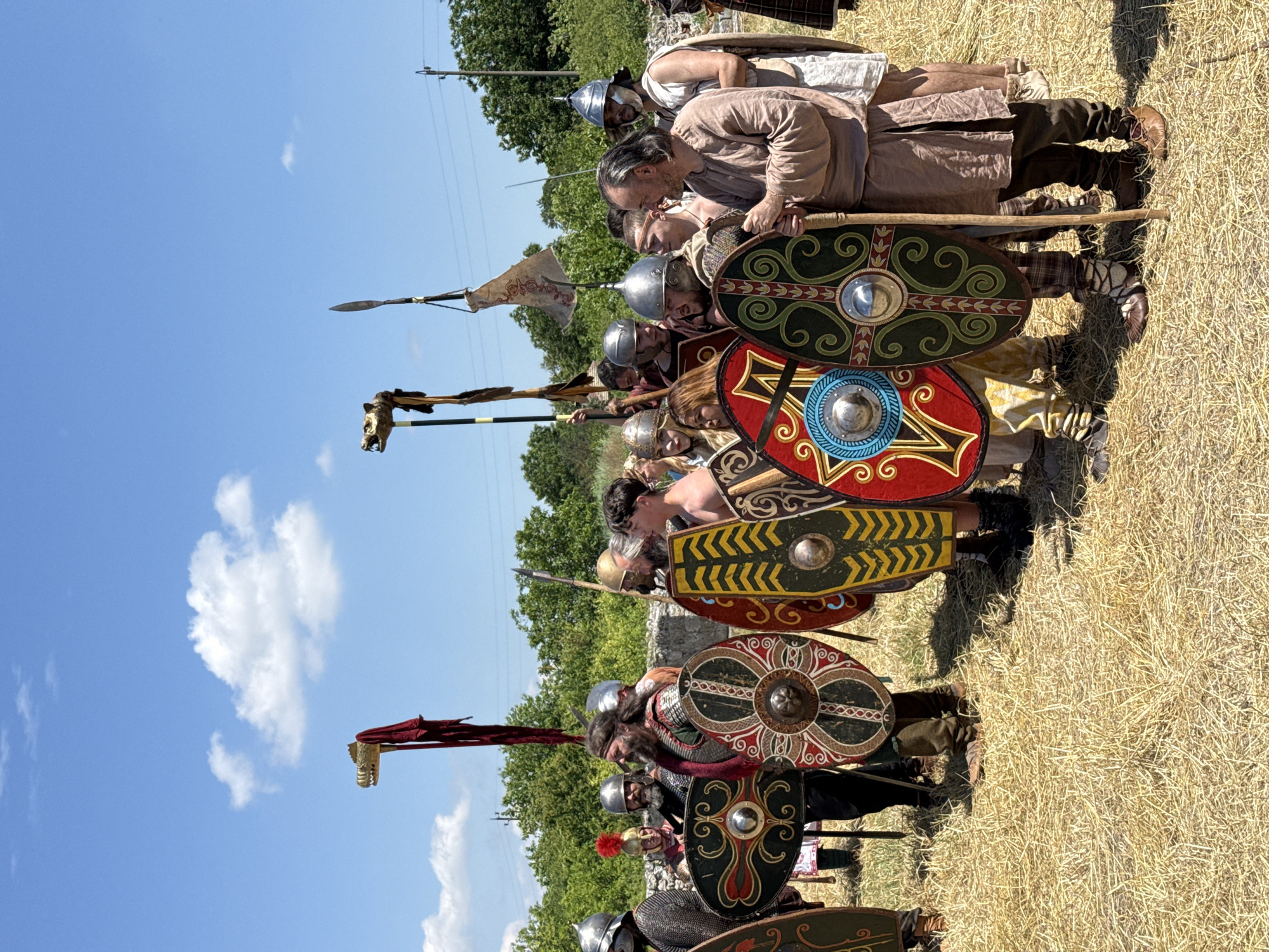 An outdoor scene in Gigen, Bulgaria, depicts a group of approximately 15-20 individuals in historical costumes, likely engaged in a reenactment. The participants are predominantly male, standing on a field covered in dry, light-brown straw under a clear blue sky with scattered white cumulus clouds.
The individuals are equipped with various pieces of period attire and gear. Many wear metal helmets of different designs, including one with a red plume on the far left-center. Clothing consists of tunics, trousers, and some chainmail, primarily in earthy tones. Several men feature long beards and hair.
Prominent objects include numerous round and oval shields. These shields are elaborately decorated with diverse geometric and organic patterns in colors such as red, green, yellow, blue, and black, each featuring a central metallic boss. Two tall standards are visible in the center-left and center-right of the upper frame; each is topped with a golden animal head, resembling a *draco* or wolf head, with one featuring a long red fabric streamer. A smaller, light-colored banner with a red design is also attached to a pole. A man in a plain brown tunic on the right holds a long wooden staff. A spear-like weapon is visible on a pole in the upper right.
The individuals are positioned in a loose formation, generally facing towards the viewer's left, with varying expressions and gazes. In the background, green trees and bushes partially obscure a low stone wall or remnants of structures. Modern electrical poles and wires are discernible in the upper right background. The scene is brightly illuminated by direct sunlight, casting short shadows.