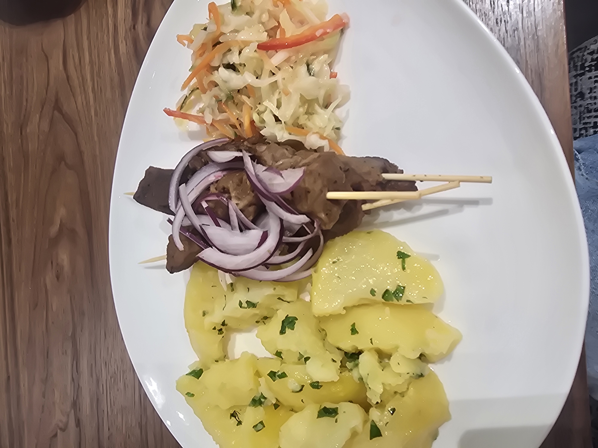 A white plate of food sits on a wooden table. The plate contains boiled potatoes with chopped parsley, a skewer with meat and red onion, and a side of coleslaw with carrots and red pepper. The food is arranged on the plate in three distinct sections. The table is made of dark wood with a visible grain. The background is out of focus but appears to be a restaurant or dining setting. There is no indication of the city of Ulan-Ude, Russia.