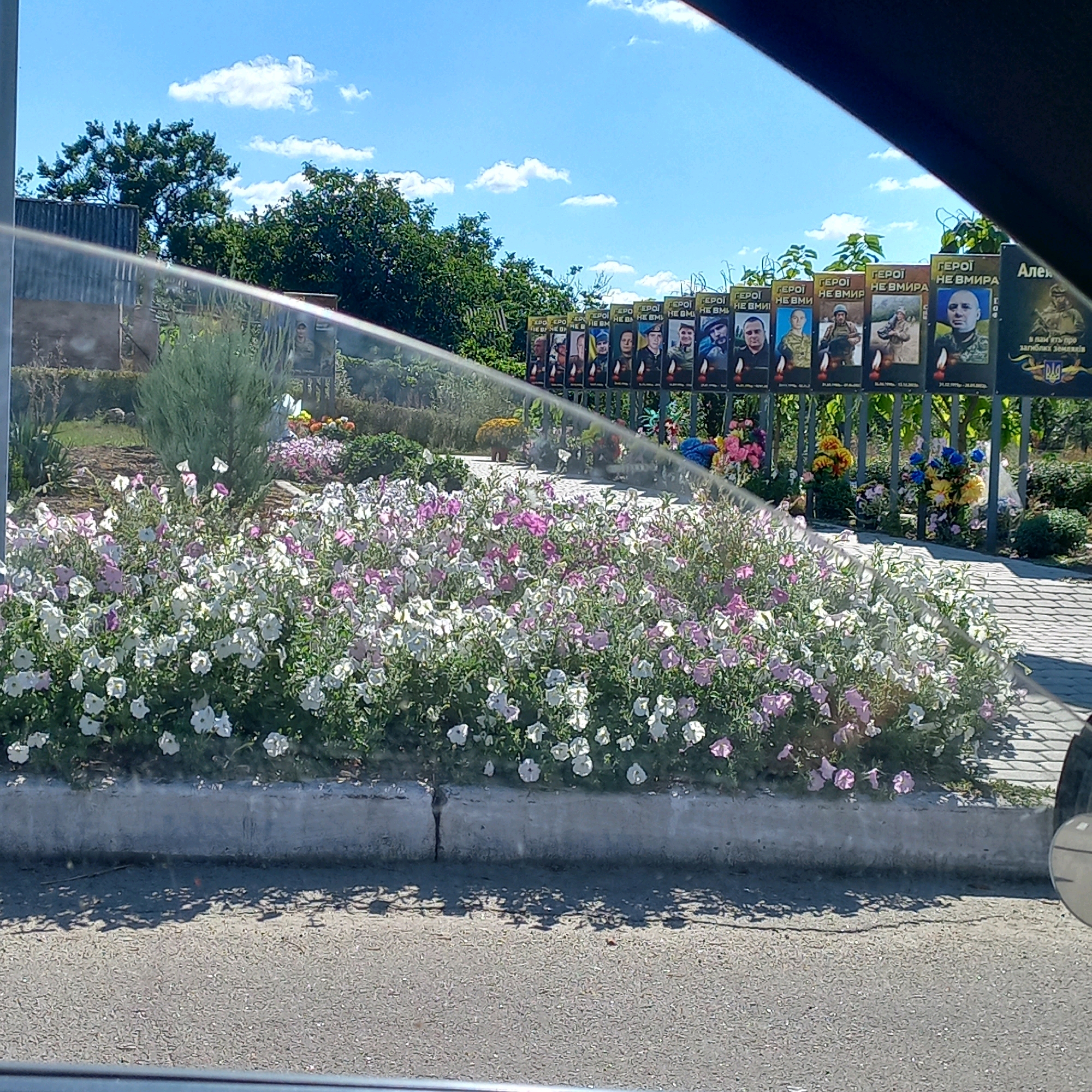 The image captures a scene featuring a floral arrangement in the foreground, with a backdrop of memorial posters of Ukrainian soldiers against a fence. A sprinkler is visible spraying water over the flowers. In the background, trees and a blue sky with scattered clouds complete the setting. The memorial posters, likely located in Klyntsi, Ukraine, display portraits of soldiers with text in Ukrainian, honoring their service. The overall atmosphere suggests a solemn yet vibrant tribute to fallen heroes, set within a public space.