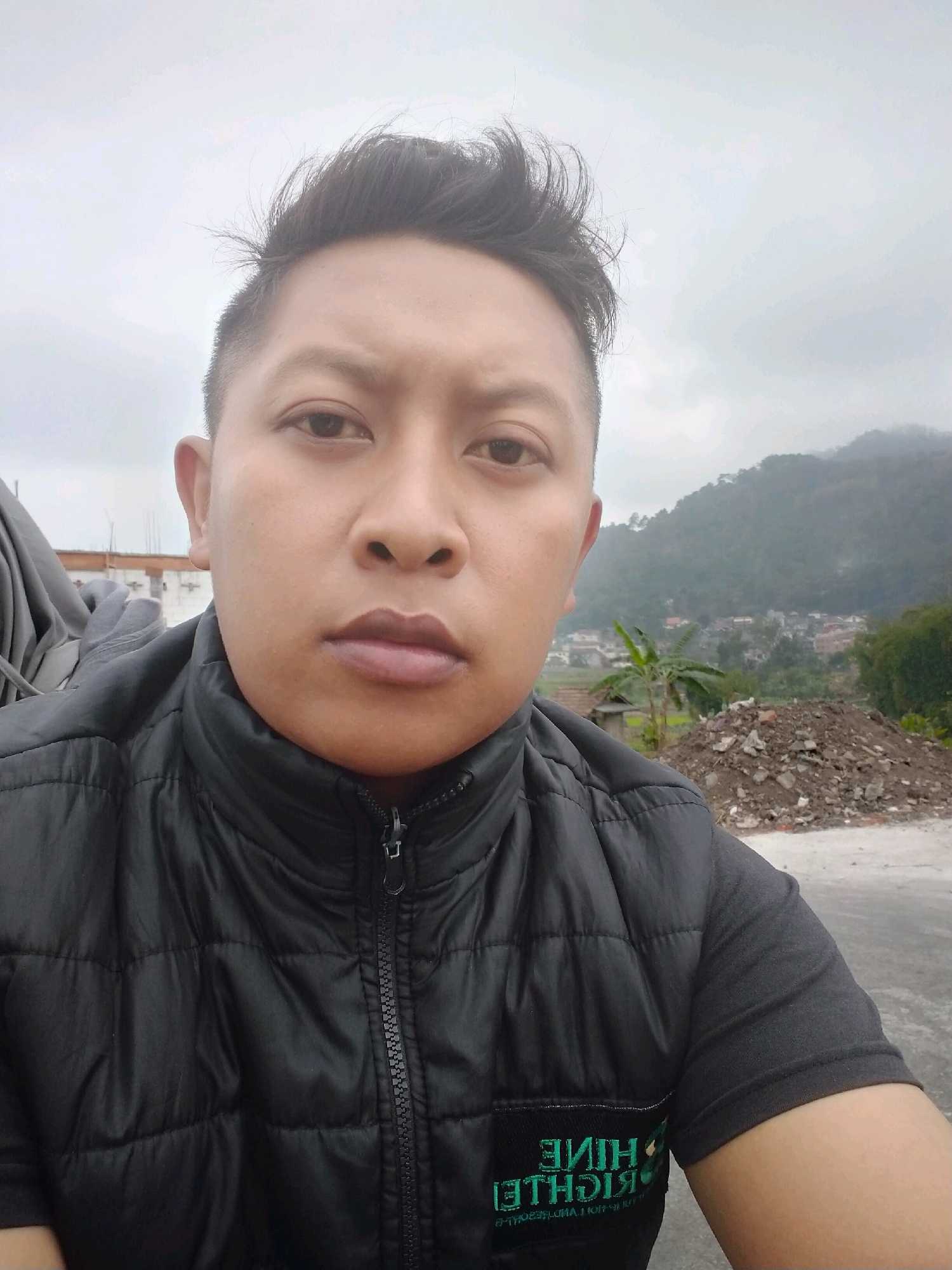 A selfie is taken by a person with short, dark hair and fair skin, wearing a black puffer vest over a dark gray t-shirt. The vest is zipped up, and the t-shirt has a green logo with white lettering. The person's face is centered in the frame, with a neutral expression. In the background, a misty, mountainous landscape is visible, with houses and vegetation scattered on the hillsides. A pile of dirt and rocks is located to the right of the person. The scene appears to be an outdoor location, possibly in a rural or semi-rural area of Batu, Indonesia, given the mountainous terrain and architecture. The sky is overcast and cloudy.