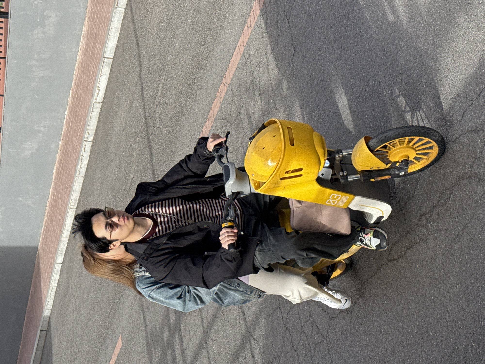 Two people are on a yellow scooter on asphalt. The person in the front is wearing glasses, a striped shirt, and a black jacket and is holding the handlebars. The person in the back is wearing a denim jacket. The background includes a curb and a building. This is likely Zhongtun, China.