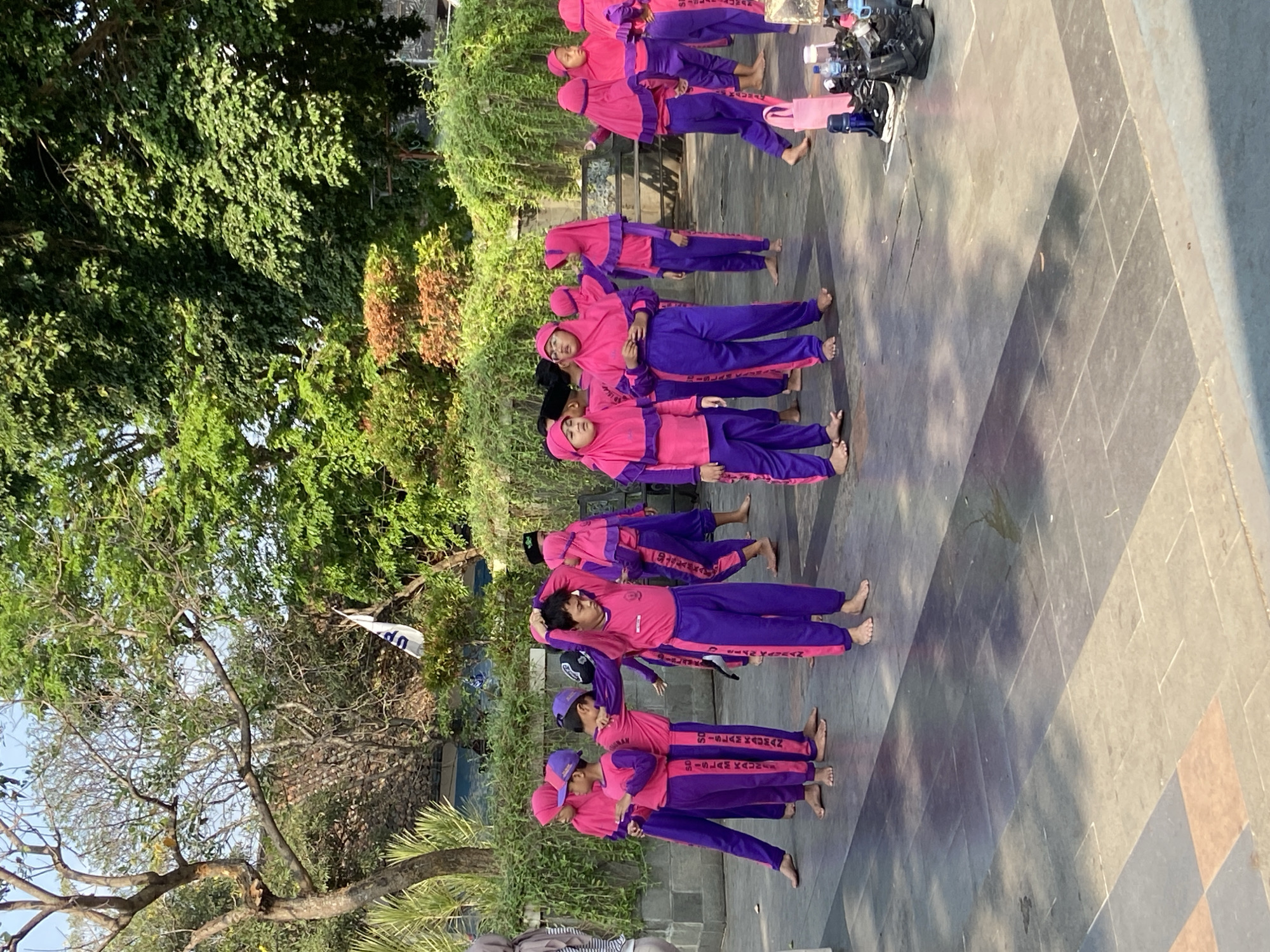 In Kecamatan Pati, Indonesia, a group of children are practicing what appears to be a dance or performance outdoors on a paved area. They are all wearing matching uniforms consisting of bright pink long-sleeved tops and purple pants, with some also wearing matching pink headscarves. The children are barefoot. The group is spread out, with some standing still, others seemingly in motion, and one child is adjusting their headscarf. Behind them, there is lush green foliage and trees, suggesting a park or courtyard setting. In the background, a white flag with blue lettering is visible, and a few structures are partially obscured by the vegetation. The lighting indicates it is daytime, with shadows cast on the pavement. Several pairs of shoes and water bottles are visible on the right side of the image, suggesting this is a performance or practice session.
