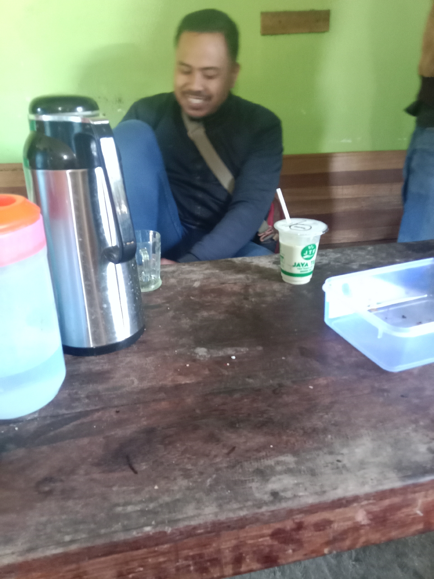 In Leksono, Indonesia, a medium shot captures a man seated at a wooden table, smiling. He wears a dark jacket and blue pants, with a tan strap across his chest. On the table, a metallic thermos, a clear glass, a cup of light-colored beverage with a straw, and a transparent plastic container are visible. The background features a light green wall and wooden paneling. The man's posture and facial expression suggest a relaxed and casual atmosphere. The setting appears to be an indoor space, possibly a cafe or a home.