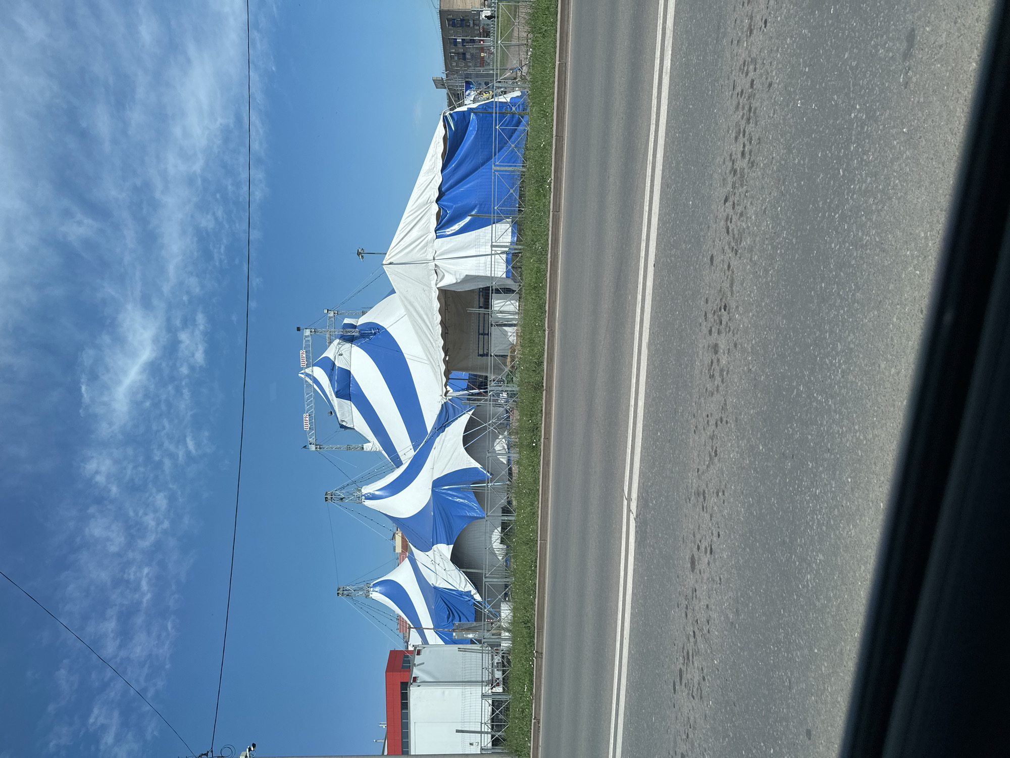 The image depicts a large, blue and white striped circus tent complex situated in a flat, open area. The tents are supported by metal scaffolding and appear to be partially erected or dismantled. In the background, there are buildings, including a red-and-white structure and a multi-story building, suggesting an urban or suburban setting. A paved road with white lane markings is in the foreground, and a grassy area with some sparse white flowers separates the road from the tents. The sky is clear blue with wispy white clouds. The overall scene is a daytime outdoor view. The image does not contain any discernible individuals or specific actions. The location is identified as Sterlitamak, Russia.