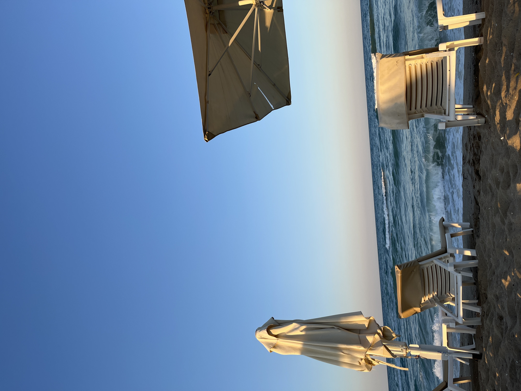 This is a wide shot, taken at a low angle, of a beach in Obzor, Bulgaria. In the foreground, there are several empty, white plastic lounge chairs on the sand. Some of these chairs are stacked, while others are laid out. A closed, light-colored beach umbrella stands on the left side of the frame, with the top of an open umbrella visible on the right. The sea is in the background, with gentle waves breaking on the shore. The sky is clear and blue, with a soft gradient near the horizon, suggesting it is either early morning or late afternoon. The sand is light brown and appears dry in most areas. There are no people visible in the image.