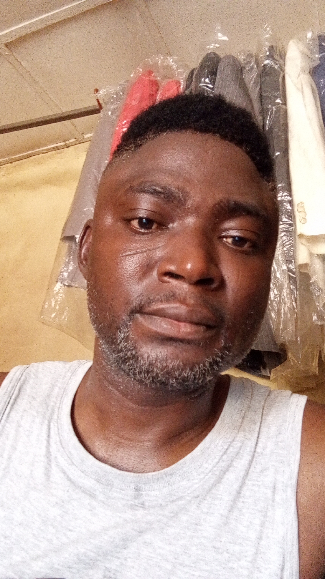 The photograph presents a close-up view of a middle-aged black man with a dark complexion. He has short, dark hair and a greyish beard. His face appears to have some texture and lines, including a possible scar near the left temple area. He is wearing a light grey tank top. Behind him, there are garments, likely suits, covered in plastic. These are suspended, presumably in a retail or storage setting. The location given, Sabon Tasha, Nigeria, suggests that this image might be of a tailor, dry cleaner, or similar business in the city. The exact address of the place is not visible.