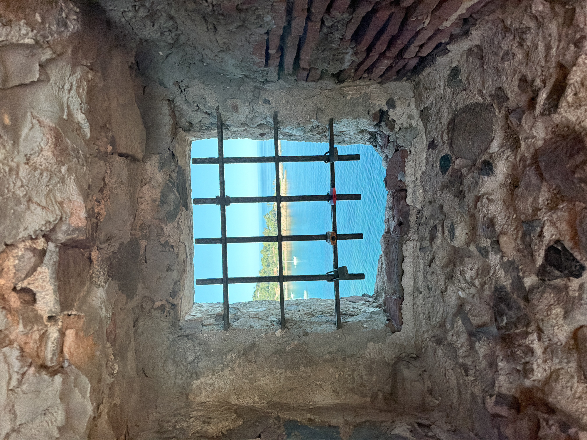 A close-up, interior view looks through a barred window of a stone structure. The window opening reveals a vibrant blue sky and a turquoise sea with several sailboats visible. In the distance, a lush green coastline with trees and buildings can be seen. The bars of the window are old and rusty, with two small padlocks attached to them, one with a red ribbon. The stone walls framing the window are rough and weathered, showing a mix of large stones and cement, with some red brickwork visible on the right side. The scene is suggestive of a historic fortification overlooking a coastal town, possibly in the Mediterranean, given the style of the architecture and the clear, bright seascape. The overall impression is one of a protected or confined vantage point offering a beautiful, expansive view. The location is identified as Tortolì, Italy.