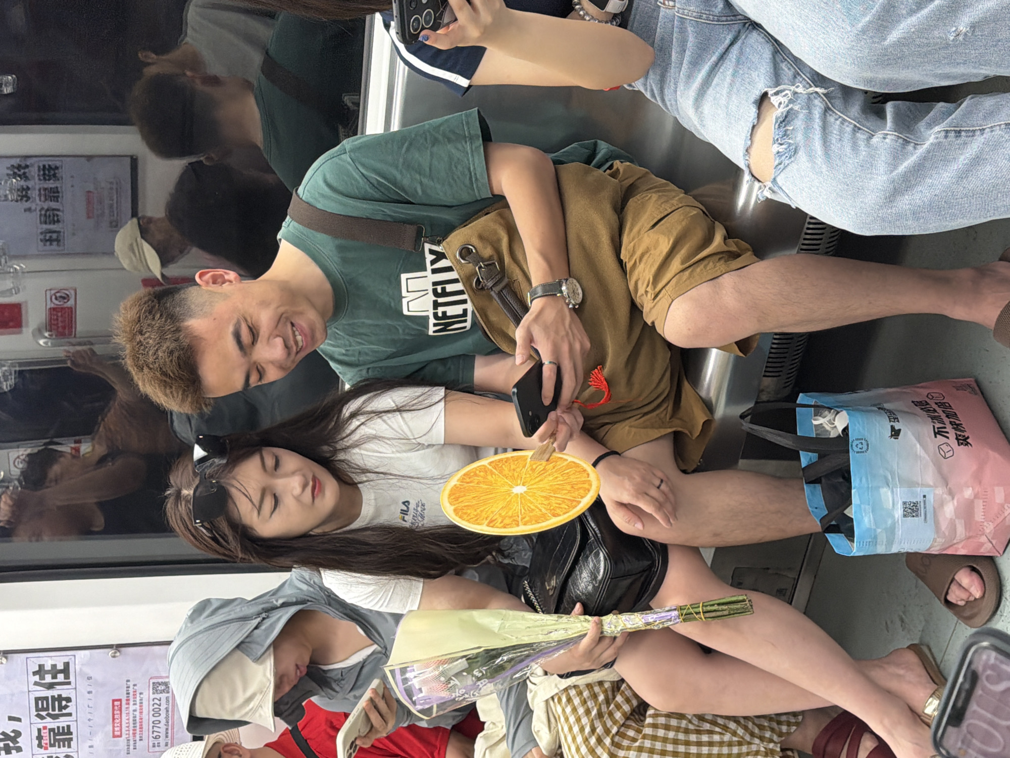 In Yuzhong District, China, on a subway train, a man and a woman are seated next to each other. The man, wearing a green t-shirt with a Netflix logo and brown shorts, is smiling and looking down at his phone, which he is holding in his left hand. A brown canvas messenger bag is slung across his right shoulder. The woman, wearing a white t-shirt and dark shorts, has sunglasses perched on her head. She is holding a bouquet of flowers in her left hand and an orange slice prop in her right hand, and is looking at her phone. They are sitting on a metal bench, with other passengers visible in the background. A patterned shopping bag is on the floor between their feet. The subway car interior is visible, with advertisements and handrails.