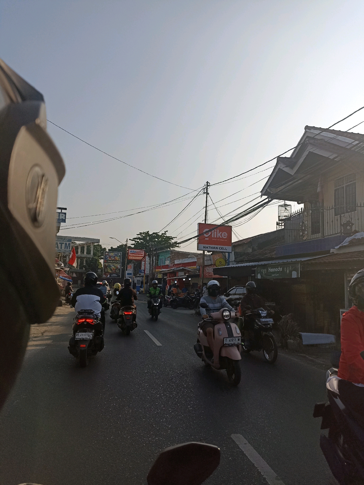 The image shows a street scene in Bandar Lampung, Indonesia, likely during daylight hours with clear skies. The perspective is from the viewpoint of someone riding a motorcycle, with a portion of a dark helmet in the foreground on the left. A line of motorcycles is proceeding down a paved road, with drivers and passengers wearing helmets. On the right side of the road, there are buildings with various signs, including "Alfamart" and "Hannochs." Further down the street, more buildings and signs are visible, including "ATM" and a red and white Indonesian flag. Power lines crisscross overhead. The overall atmosphere is that of a typical busy street in an Indonesian city.