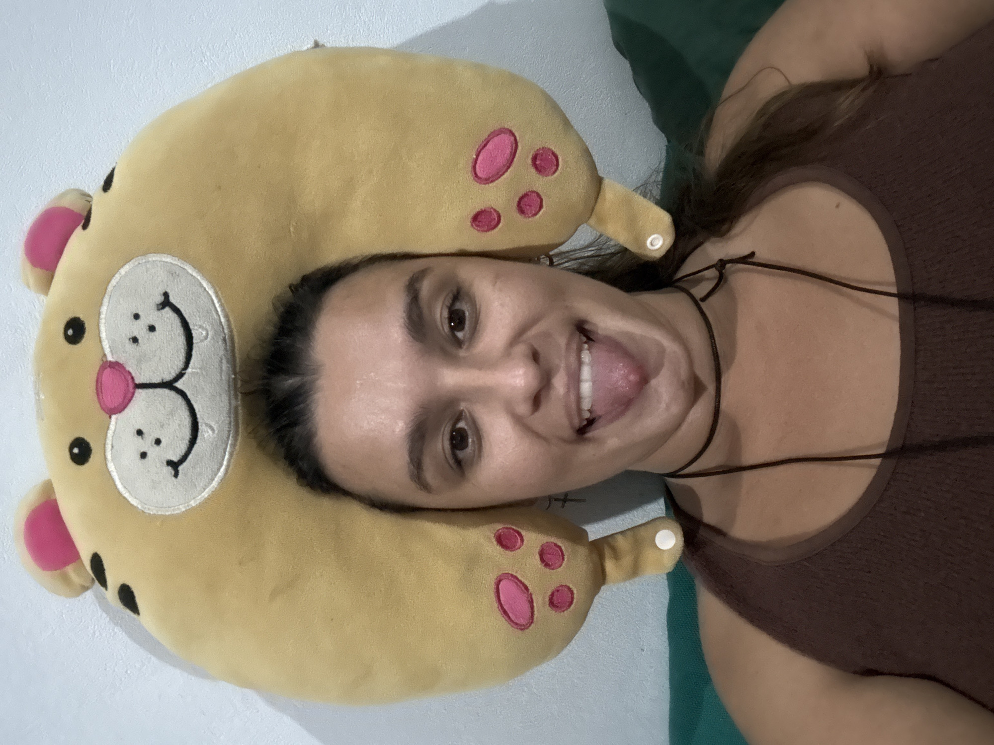 A woman lies on a surface, head resting on a yellow, crescent-shaped pillow with pink paw prints. She sticks her tongue out and wears a brown tank top with a black necklace. The background is a light blue wall and a green surface. The location is Pejeng, Indonesia.