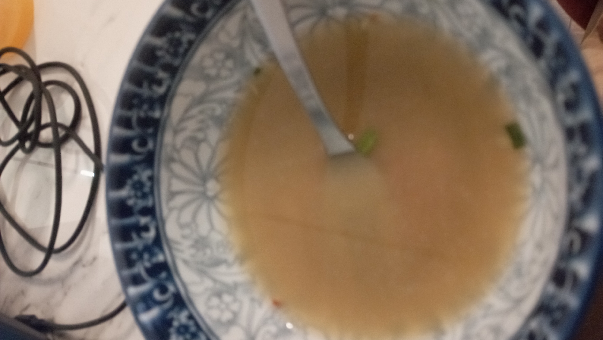 A close-up, slightly blurred image depicts a bowl of broth with a spoon resting inside. The bowl has a blue and white patterned design. Small green pieces, likely scallions, are scattered in the broth. To the left of the bowl is a tangled black cord on a light-colored surface, possibly a countertop. A blurred, out-of-focus object is in the upper left corner. The context of Nha Trang Ward, Vietnam is provided but does not offer any visible evidence in the image.