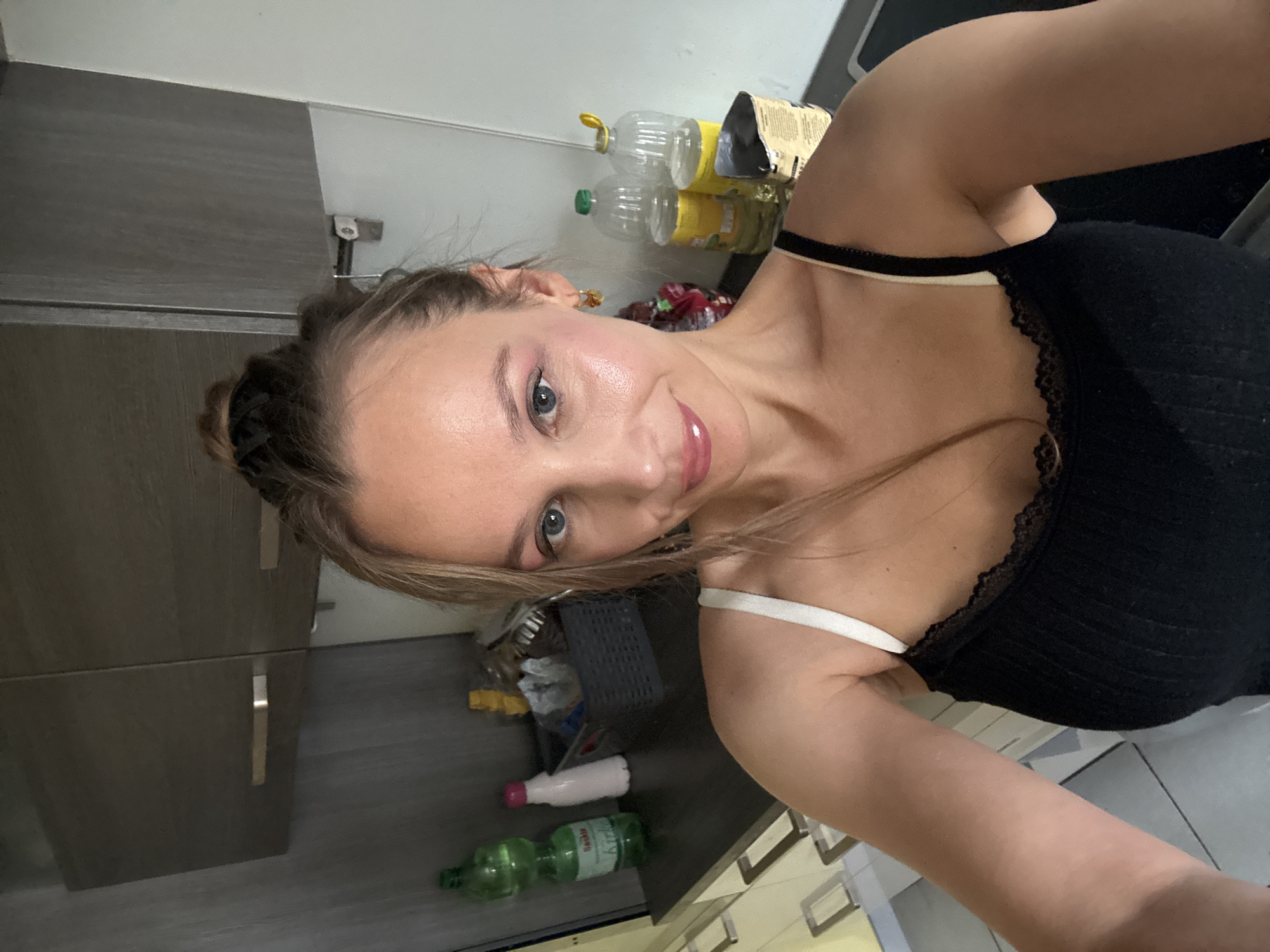 A woman is taking a selfie in a kitchen in Überherrn, Germany. She has her hair up in a bun and is wearing a black tank top with white straps. She has on makeup, including eyeshadow and lipstick. The kitchen has grey cabinets and countertops. There are bottles of water and oil on a shelf. The woman is smiling and looking at the camera.