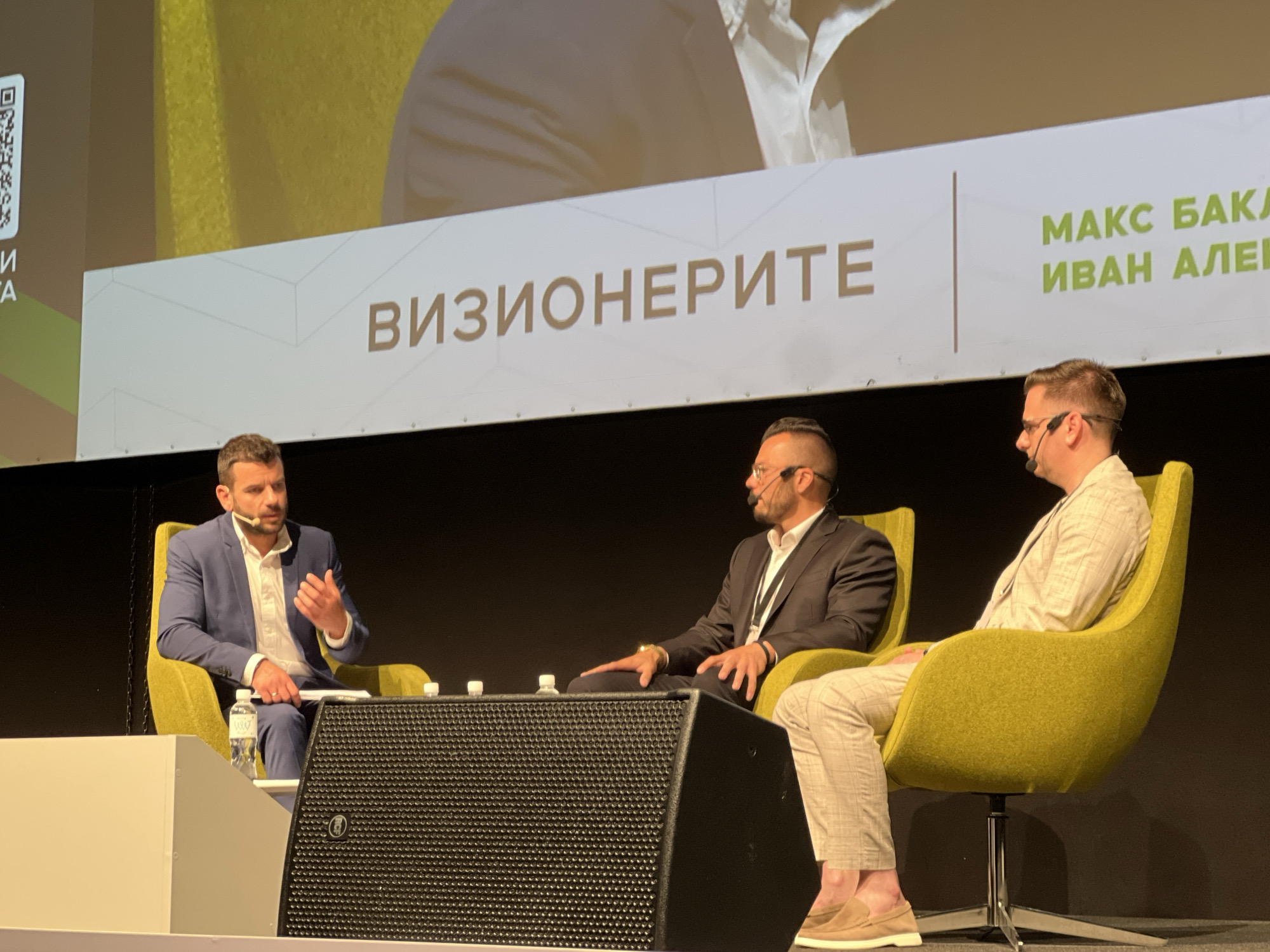 The image depicts three men seated on chairs, seemingly engaged in a panel discussion on a stage. The panel is in front of a banner that reads “ВИЗИОНЕРИТЕ МАКС БАКА ИВАН АЛЕК” which translates to “VISIONARIES MAX BAKA IVAN ALEK”. Each man is wearing professional attire. The first man, on the left, wears a blue suit. The second man, in the middle, wears a dark suit. The third man, on the right, wears a light colored suit with matching pants and loafers. All three men are wearing microphones. There are water bottles on the table between them. A speaker is placed in front of the table. The setting appears to be a conference or event, likely held indoors. Based on the writing in the banner, the event took place in Sofia, Bulgaria.