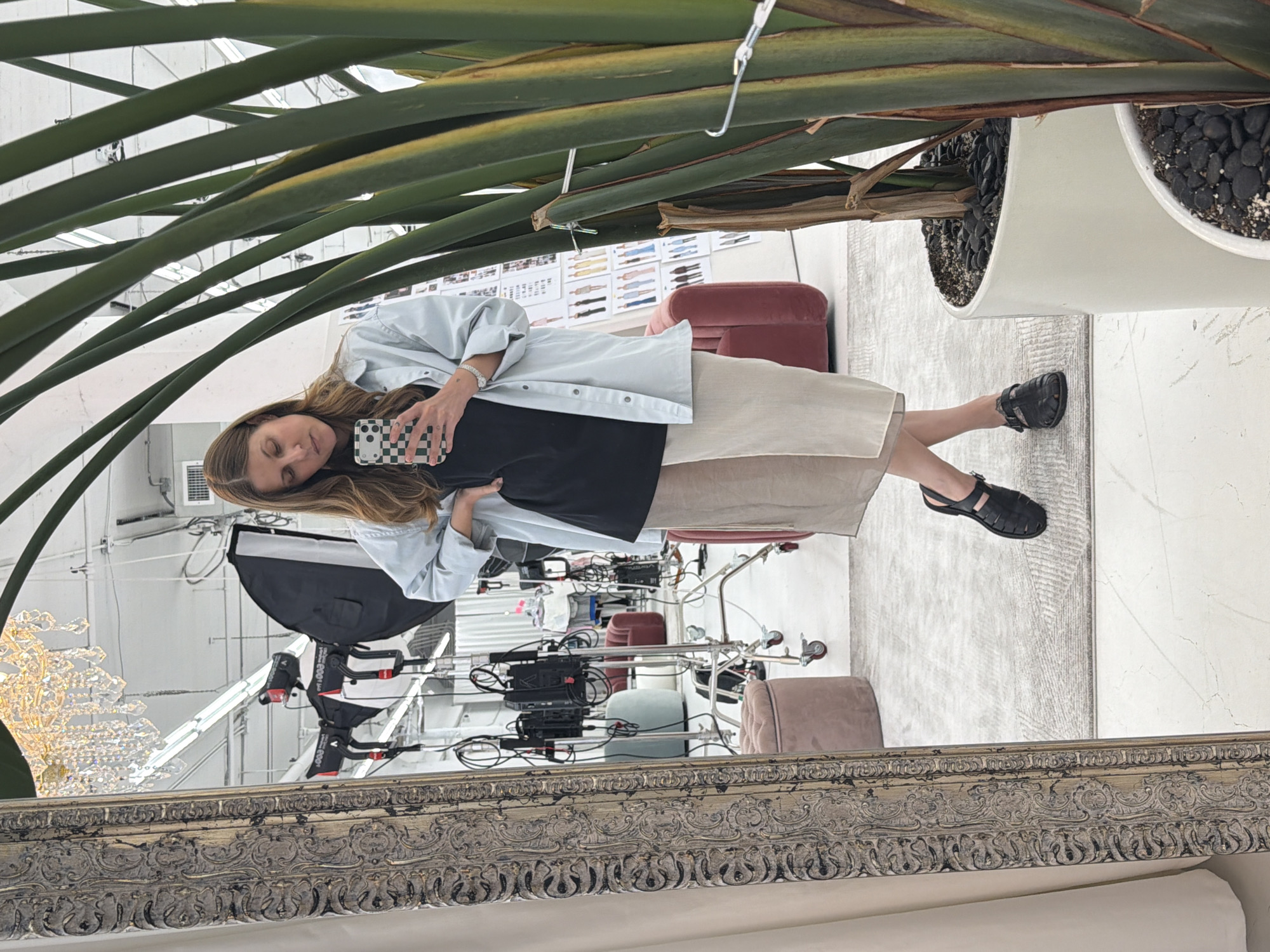 The image appears to be a reflection in a mirror, capturing a woman and aspects of what seems to be a studio or showroom. The woman is holding a phone with a checkerboard case and is wearing a blue shirt over a black top and a long beige skirt. A large plant with elongated green leaves is prominently featured in the foreground, obscuring parts of the scene. The reflection reveals part of a studio setting with equipment such as studio lights and camera stands. There are also what appear to be mood boards or design sketches on the wall. The furniture visible includes red chairs and a pink ottoman. The scene seems to be located in Los Angeles, United States, based on the style of the environment and the architectural elements visible.