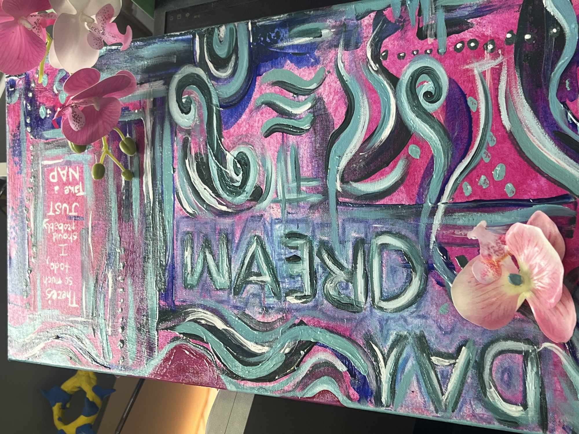 The image shows a close-up of a colorful, textured canvas with painted text and artificial orchid flowers attached. The text on the canvas appears to read "DREAM" and also includes the phrase "There's so much to do, I should probably JUST TAKE A NAP." The painting is a mix of vibrant pinks, blues, and teals, with a swirling, abstract pattern. The artificial orchids are pink and white, and are positioned in the foreground, partially obscuring the painting. The background is blurred, but appears to be an indoor setting with some objects visible. The location is not identifiable from the image.