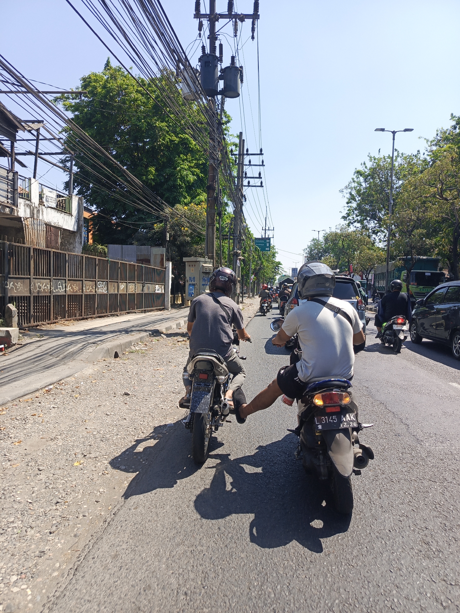 The image depicts a street scene in Calukan RT 1 RW 5 Keboansikep, Indonesia, on a clear, sunny day. The perspective is from behind two motorcyclists traveling on a paved road. The road is lined with a rough, gravel shoulder on the left. Overhead, a dense network of electrical wires and utility poles dominate the sky, with two large transformers visible on one of the poles. To the left, a weathered, rusty metal gate with graffiti runs along the side of the road, with a building partially visible behind it.
In the foreground, the two motorcyclists are prominent. The rider in front, viewed from behind, wears a gray t-shirt and a dark helmet. Their left leg is extended backward, resting on the rear of their motorcycle. The second motorcyclist, also seen from behind, wears a white t-shirt, black shorts, and a gray helmet with a black strap. This rider's license plate reads "L 3145 AAK 12.26". Both motorcycles are facing forward, indicating they are in motion or stopped in traffic.
Further down the road, a moderate amount of traffic is visible, including other motorcycles and cars, suggesting a busy urban environment. Green trees line the right side of the road, interspersed with streetlights and more utility poles. The overall impression is of a typical Indonesian street scene with abundant overhead wiring and active road traffic.