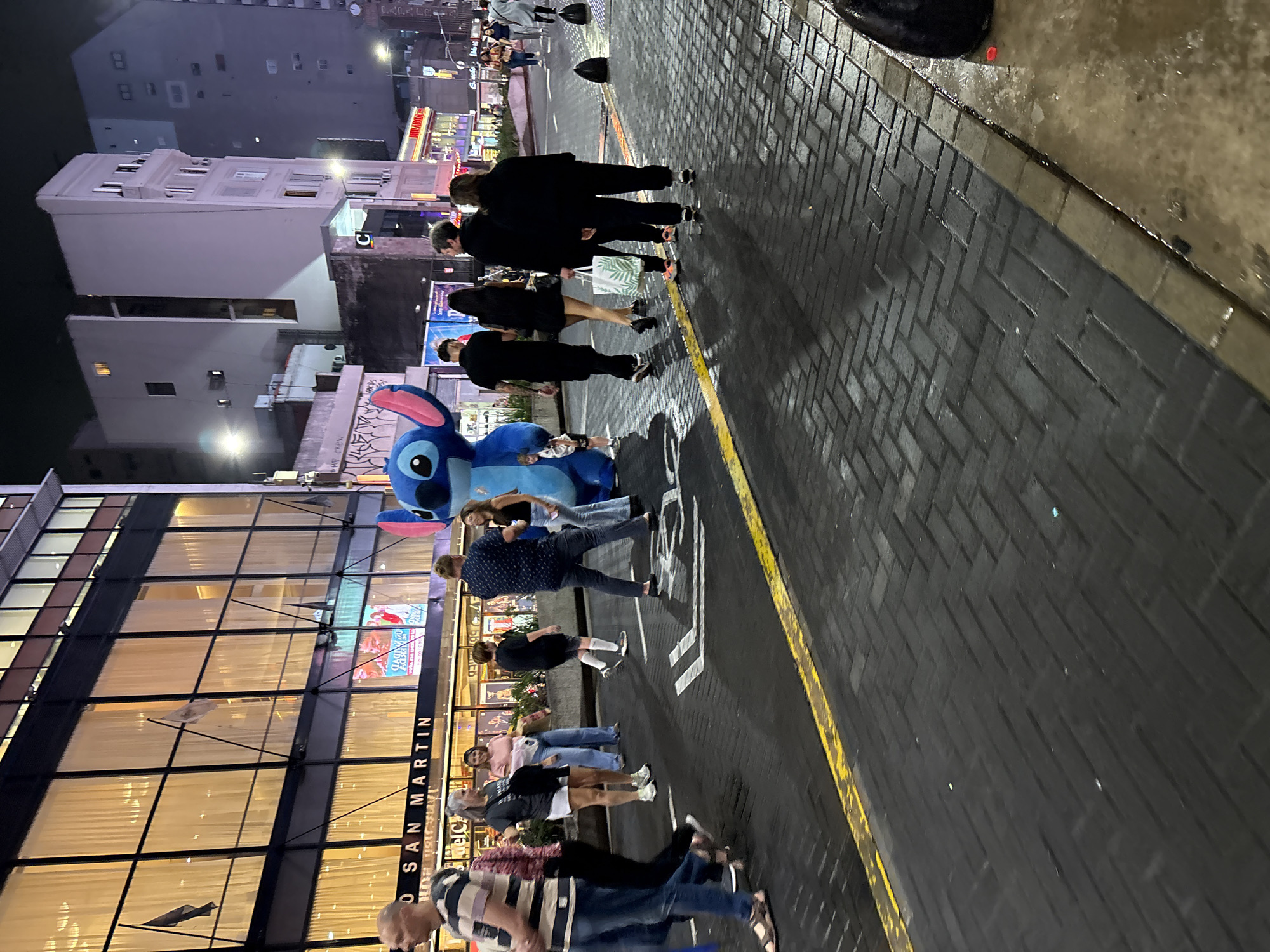 This nocturnal image depicts a bustling pedestrian area in Buenos Aires, Argentina, specifically outside the Teatro General San Martín on a dark, wet-looking paved street.
In the center of the frame, a person in a full-body blue Stitch character costume, with prominent pink ears, stands facing right, appearing to interact with nearby pedestrians. A small child is visible near the costume's left side. Several individuals are in close proximity to the Stitch character, some observing or engaging. To the immediate left of Stitch, a man in a patterned shirt, a woman in a light blue shirt, and another person are engaged in conversation.
On the left side of the street, a multi-story building identified by the illuminated sign "O SAN MARTIN" (referring to Teatro San Martín) dominates. This building features numerous large, vertically rectangular windows with light-colored curtains and is adorned with flags—two black and one white—projecting from its facade.
The street itself is paved with dark, possibly wet, interlocking pavers arranged in a herringbone pattern, marked by a faded yellow line in the foreground. Numerous pedestrians, primarily adults, are visible walking in various directions across the scene. Their attire is casual, including jeans, t-shirts, and light jackets. Further down the street, additional commercial buildings with illuminated signs, including one that appears to read "MILANESA," line the street. Streetlights provide general illumination. Black bollards are spaced along the curb in the mid-ground. Debris, including a black plastic bag, is visible on the sidewalk at the right edge of the frame.